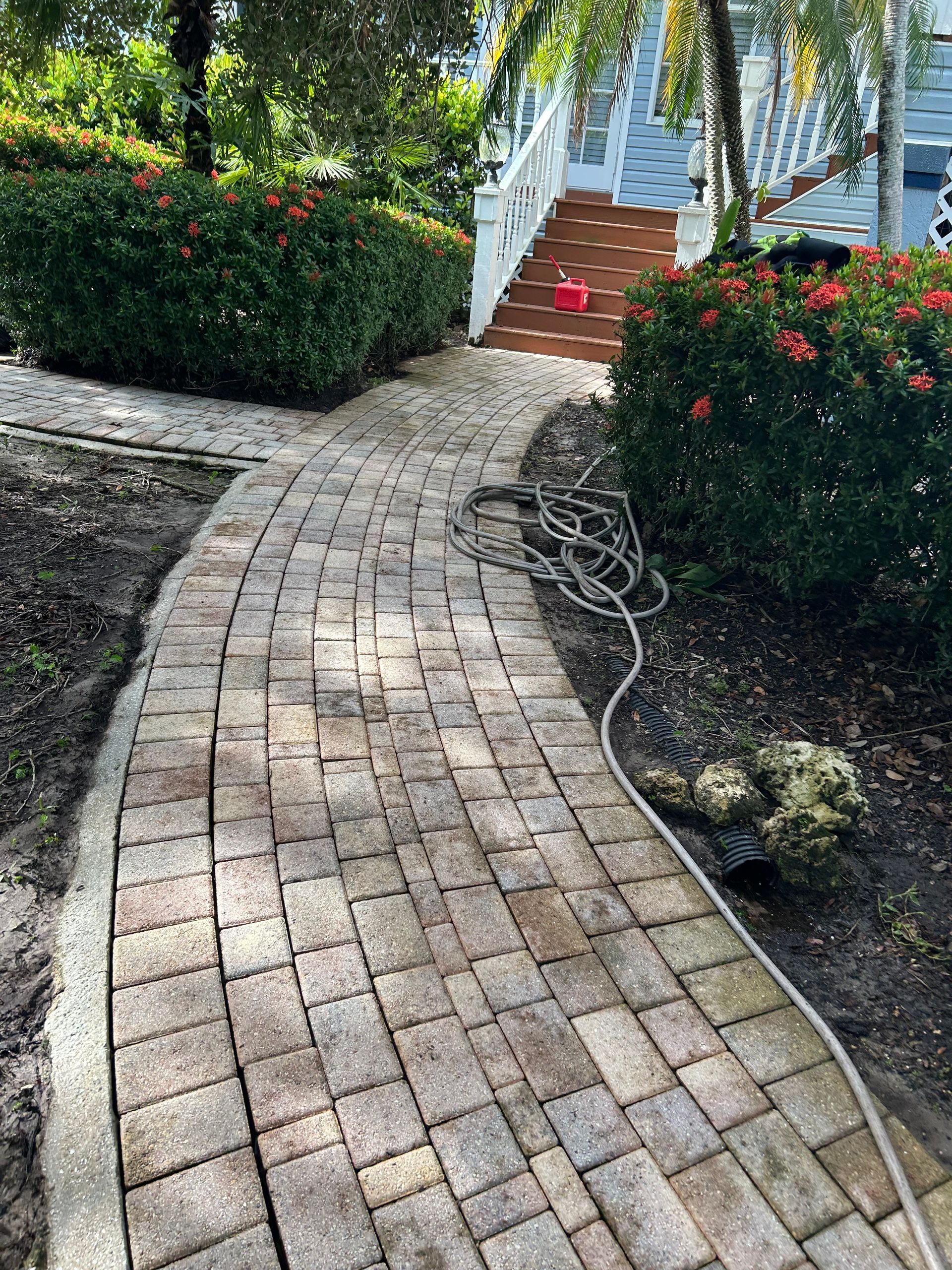 A curved brick walkway leads through a garden toward a wooden staircase and a white railing in a sunny outdoor setting.