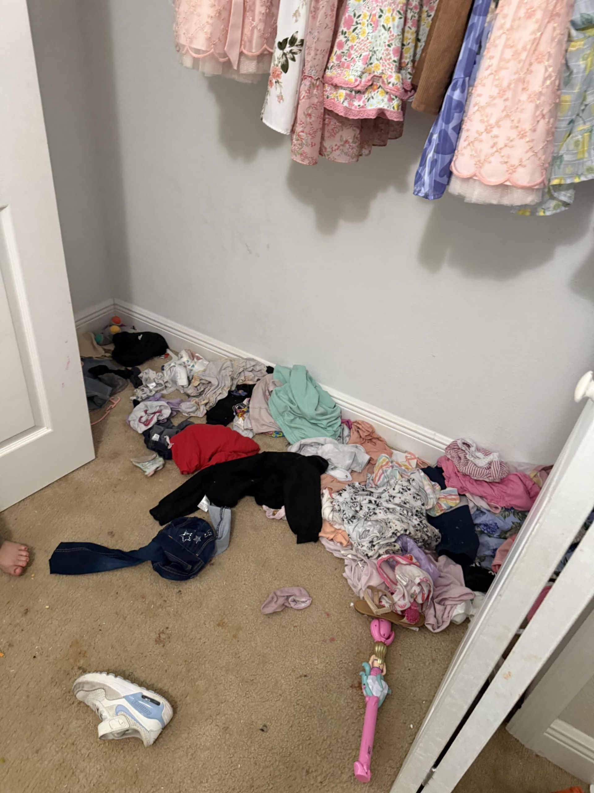 A closet floor covered in scattered clothing, a single shoe, and a toy doll, with more clothes hanging on the rack above.