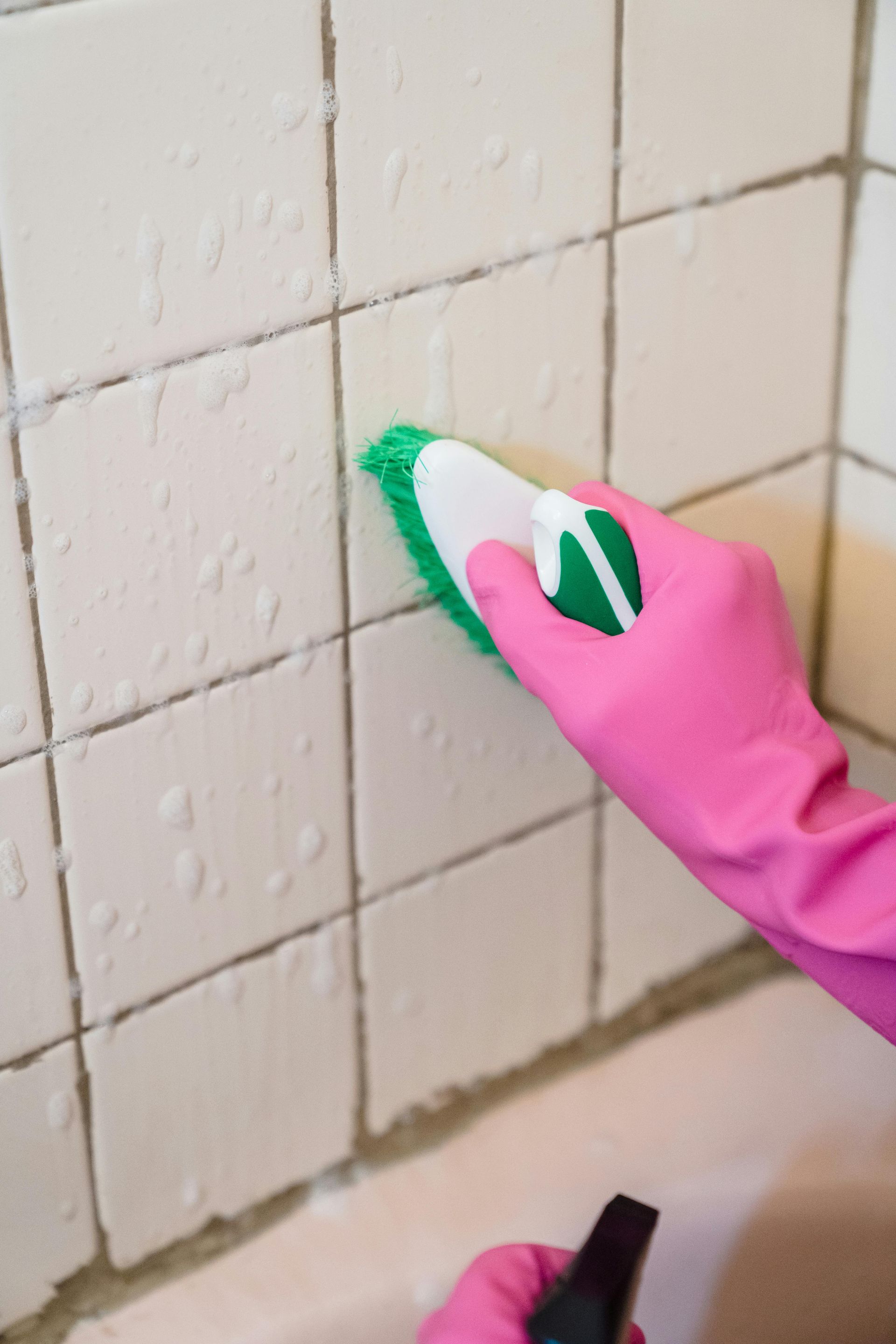 Person wearing pink gloves scrubbing white bathroom tiles with a green and white brush.