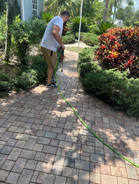 A person in a white shirt and tan shorts uses a green hose to spray a brick pathway in a landscaped garden.