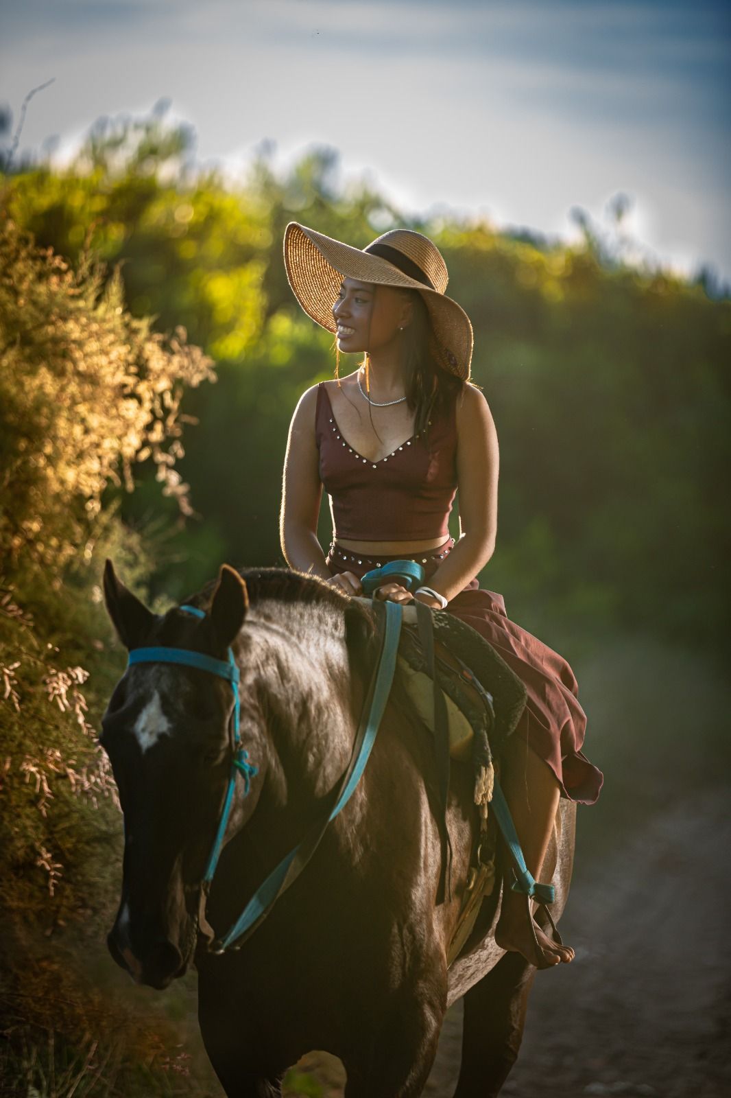 Mujer a caballo con sombrero, cabalgando por un camino de tierra con vegetación, bajo la luz del sol.