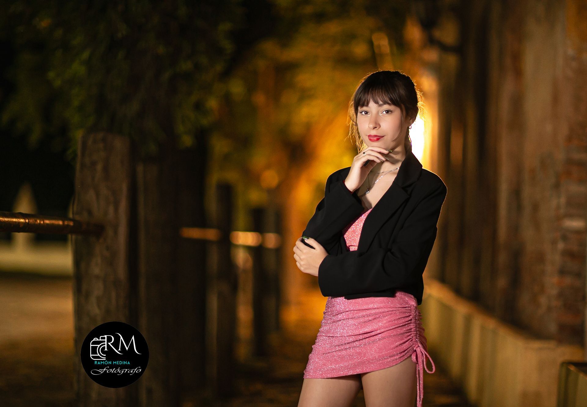 Una mujer con un vestido rosa y un blazer negro está al aire libre por la noche con la mano en la barbilla.