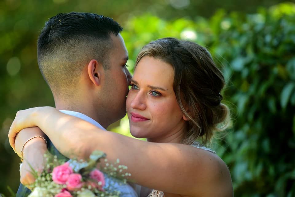 Novios se abrazan al aire libre; el novio le susurra al oído. Flores rosas visibles sobre fondo verde.