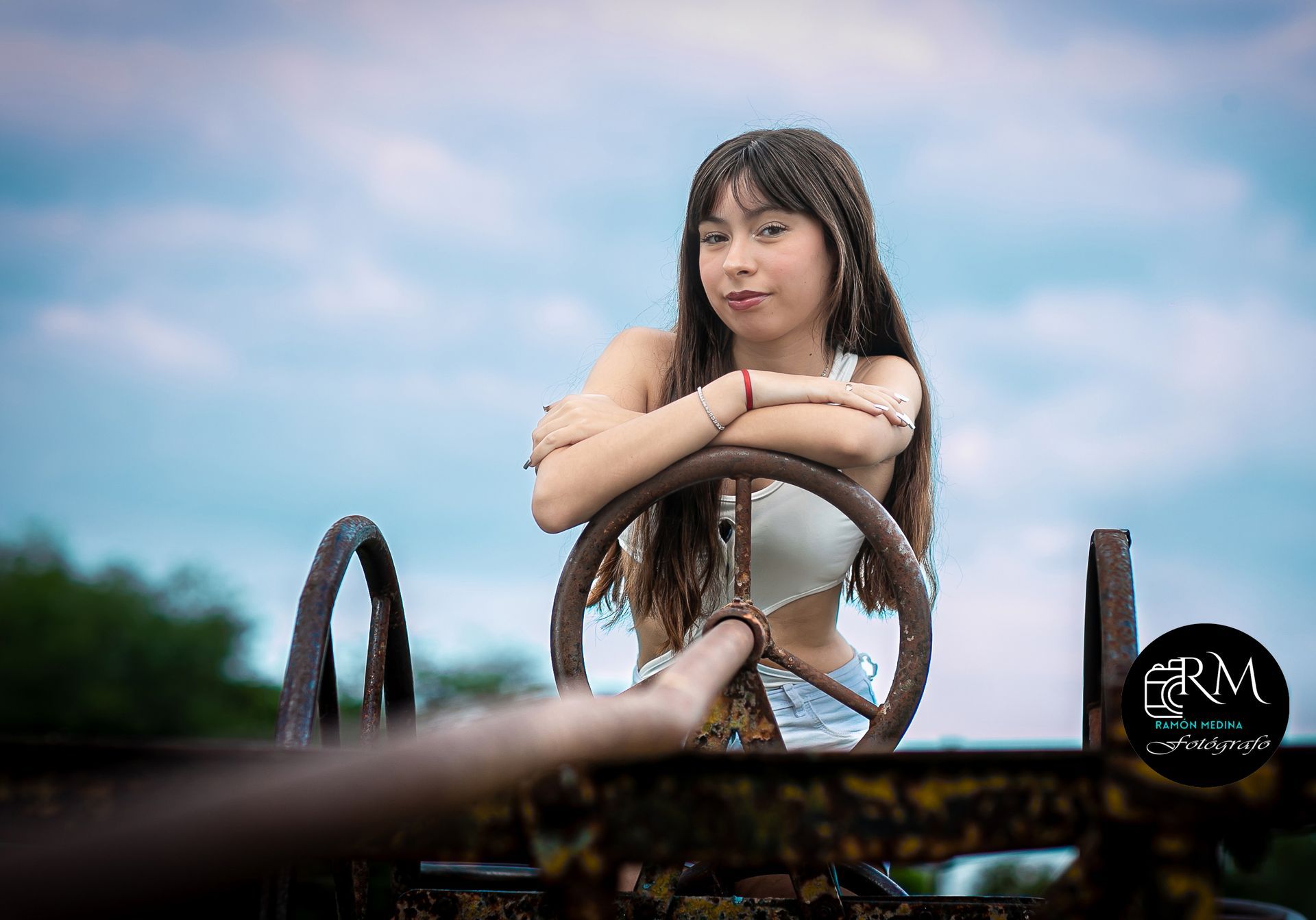 Mujer apoyada en una rueda oxidada al aire libre, con los brazos cruzados, sonriendo levemente, contra un cielo nublado.