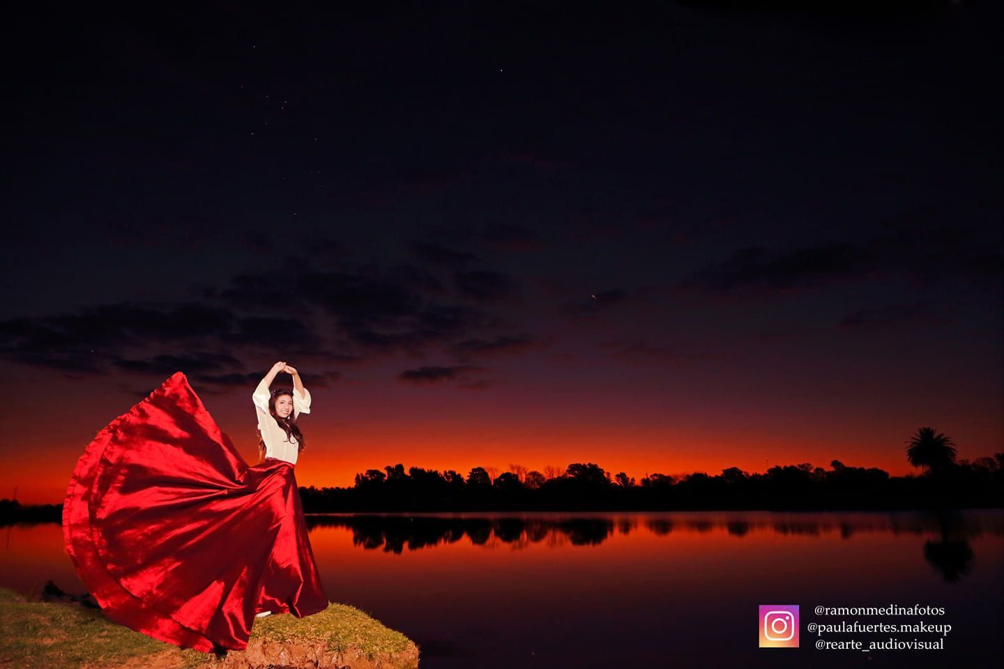Mujer con vestido rojo junto al lago al atardecer, brazos levantados, vestido suelto.