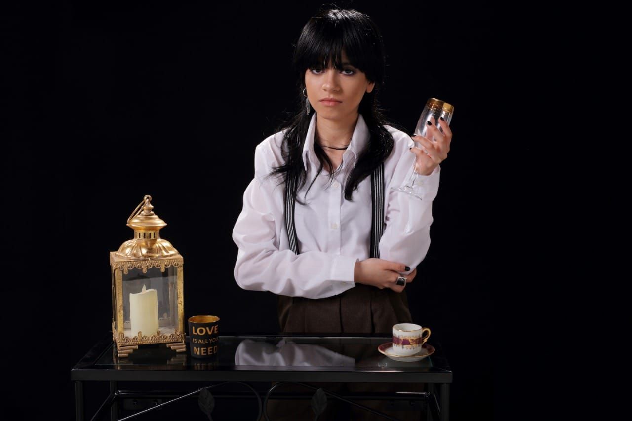 Mujer sosteniendo un vaso, mirándolo con expresión pensativa. Fondo negro, mesa con vela y taza de té.