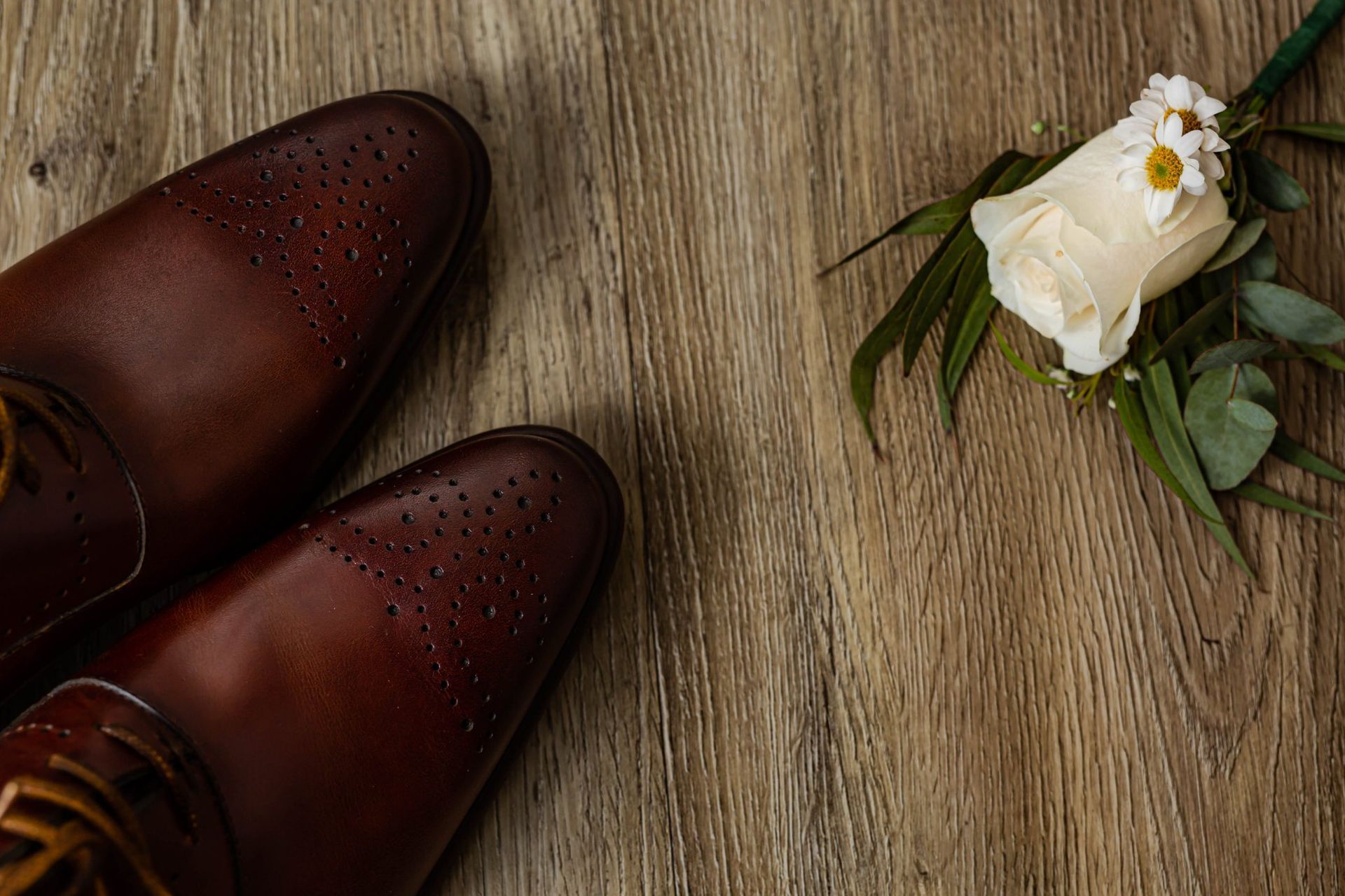 Zapatos de vestir de cuero marrón y boutonniere de rosa blanca sobre una superficie de madera.