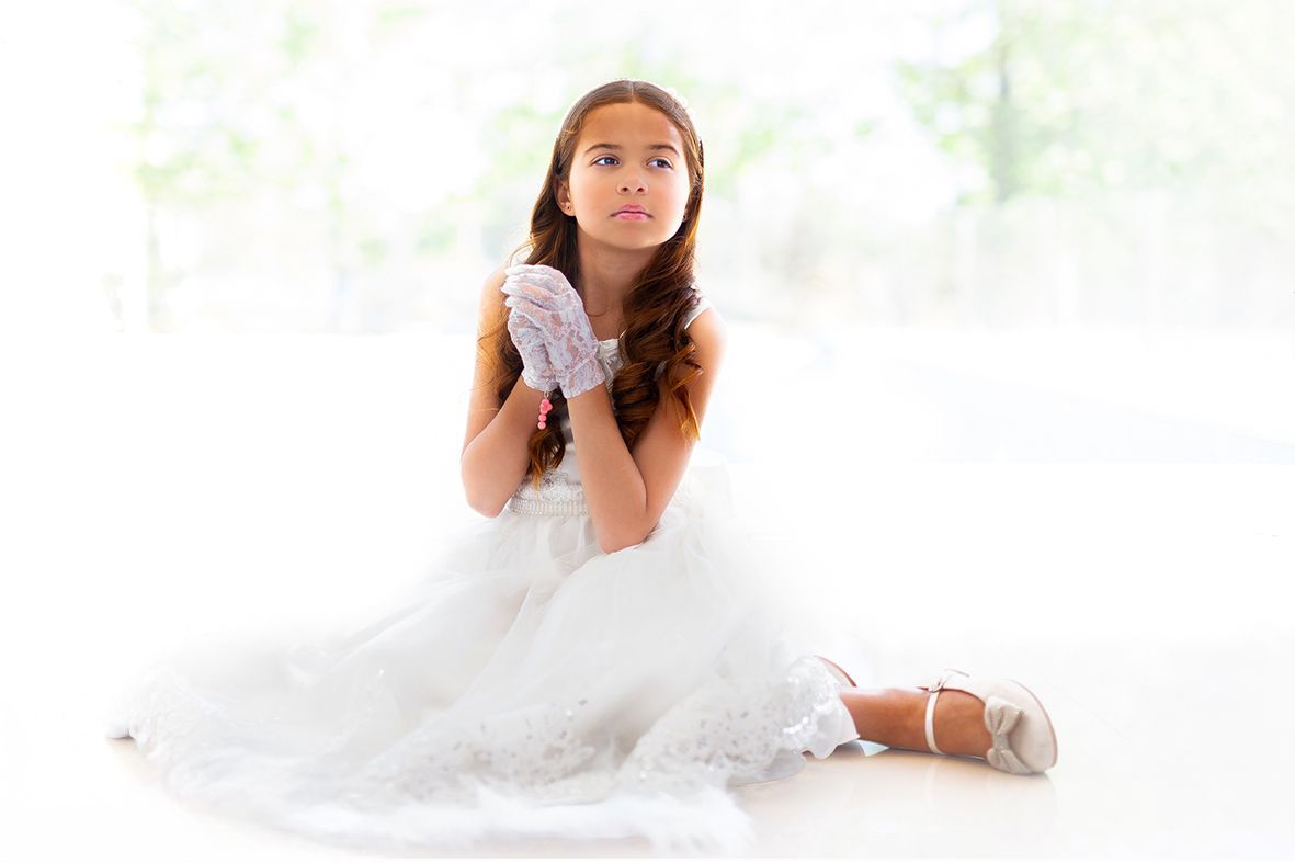 Una chica con vestido blanco se arrodilla, con las manos juntas, mirando hacia otro lado. Enfoque suave y fondo brillante.