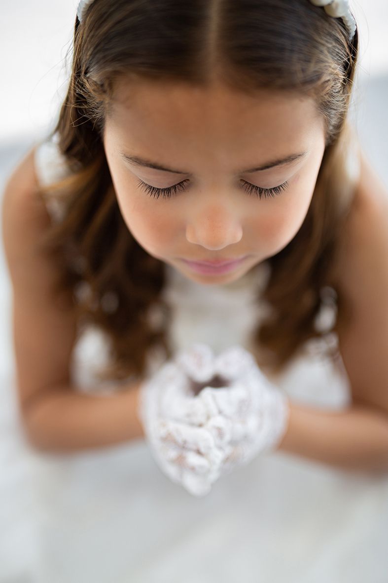 Niña con vestido blanco y guantes, ojos cerrados, manos entrelazadas, cuentas en las manos.