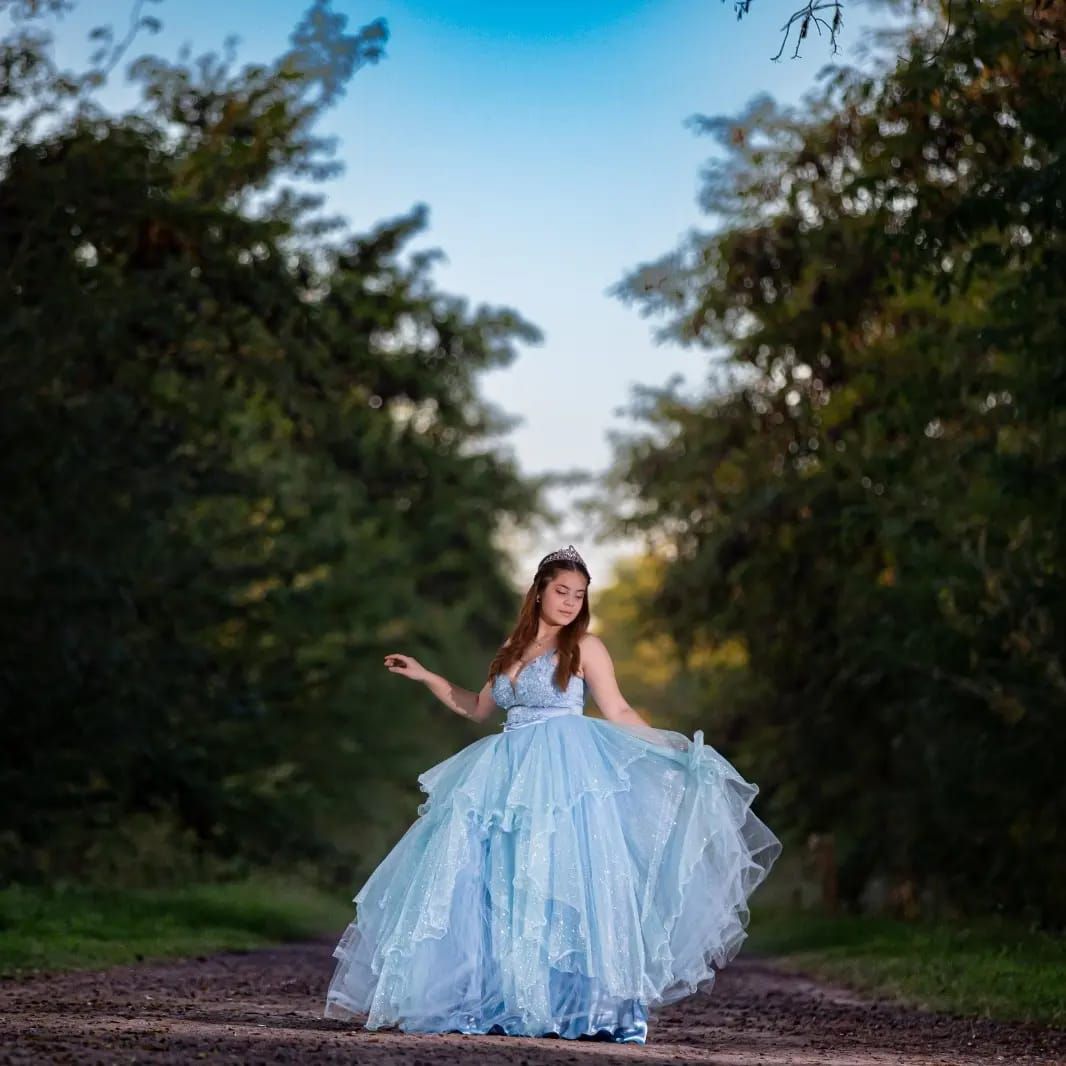 Una mujer con un vestido azul está parada en un camino de tierra en un bosque, con los brazos extendidos y sonriendo.