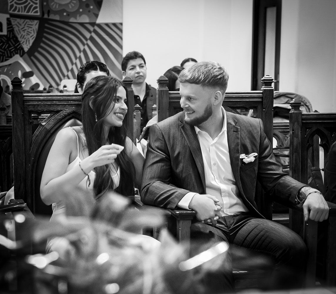 Una pareja sonriéndose, sentada en bancos de madera. El hombre con blazer y la mujer con vestido.