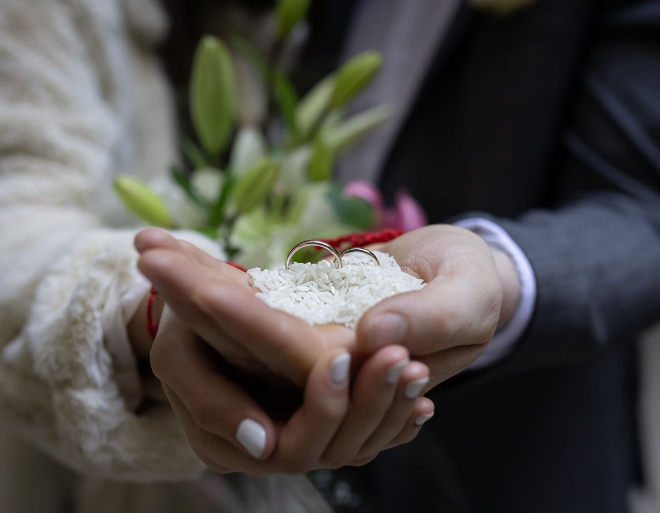 Manos sosteniendo arroz blanco con dos anillos de boda, flores borrosas en el fondo.