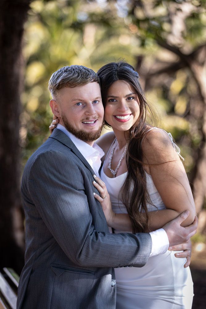 Una pareja sonriente abrazándose al aire libre. El hombre lleva traje y la mujer vestido blanco.