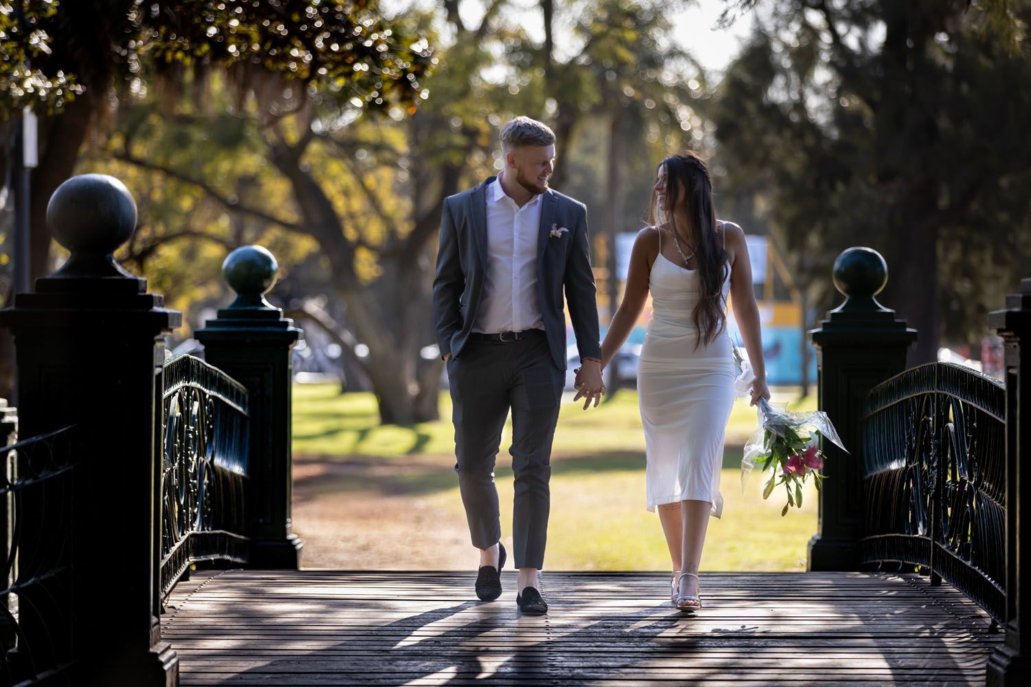 Una pareja vestida de boda camina de la mano sobre un puente, sosteniendo flores, fondo de un parque soleado.
