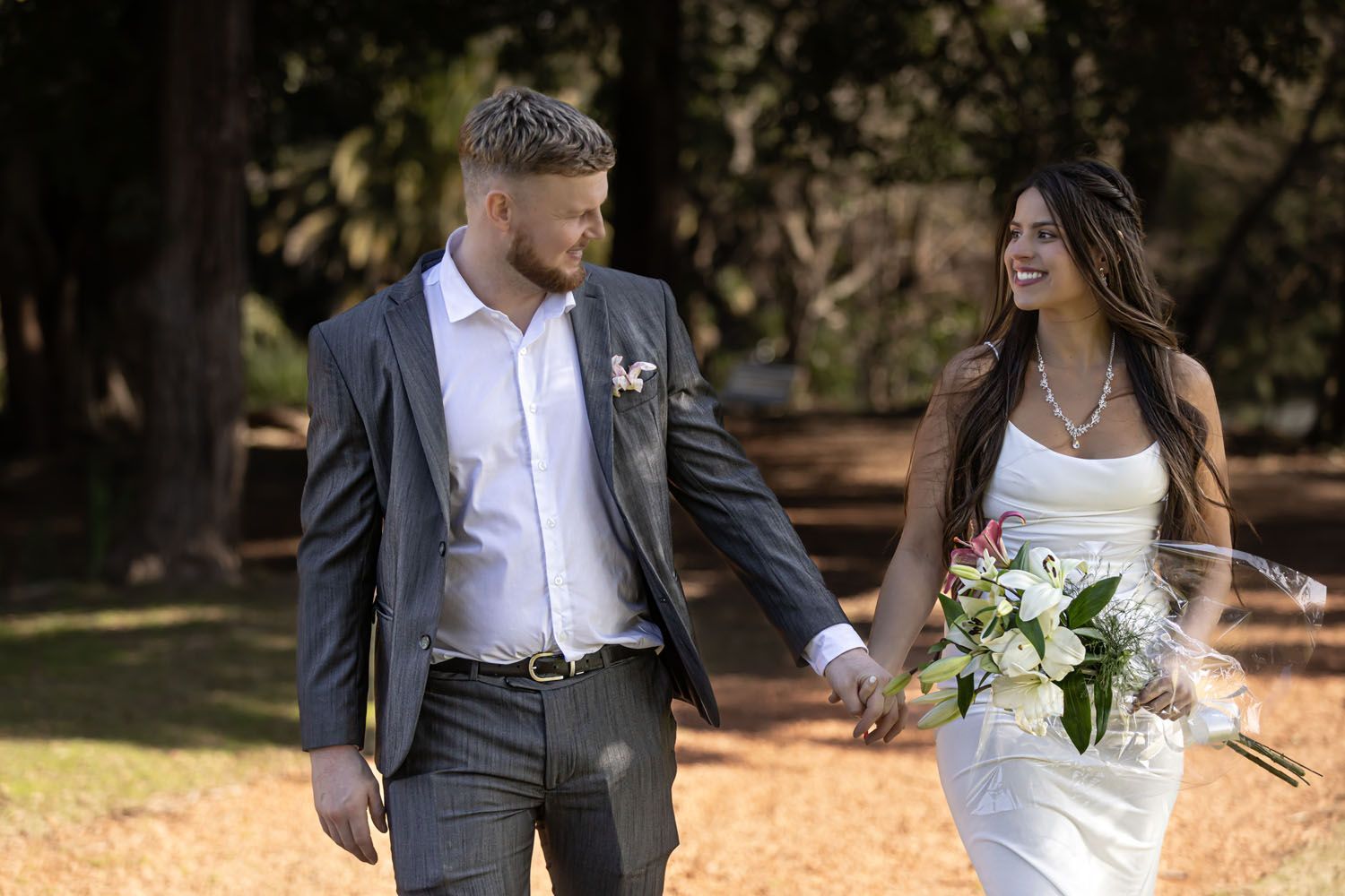 Novios caminando de la mano por un parque, sonriendo. La novia lleva un ramo. Luz del sol.
