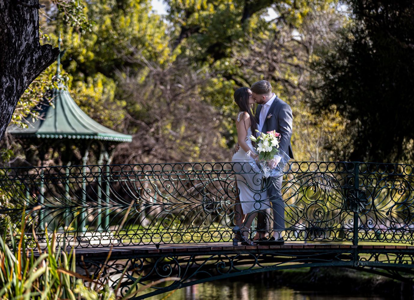 Una pareja besándose en un puente decorativo en un parque. Árboles y un cenador al fondo.