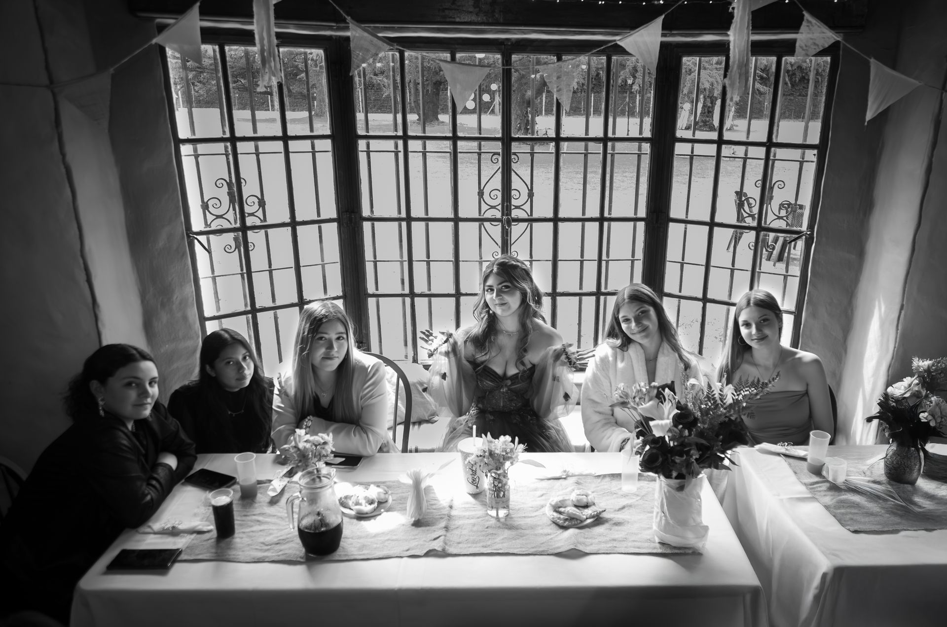 Seis personas sentadas a una mesa frente a una ventana. Hay flores y bebidas sobre la mesa. Blanco y negro.