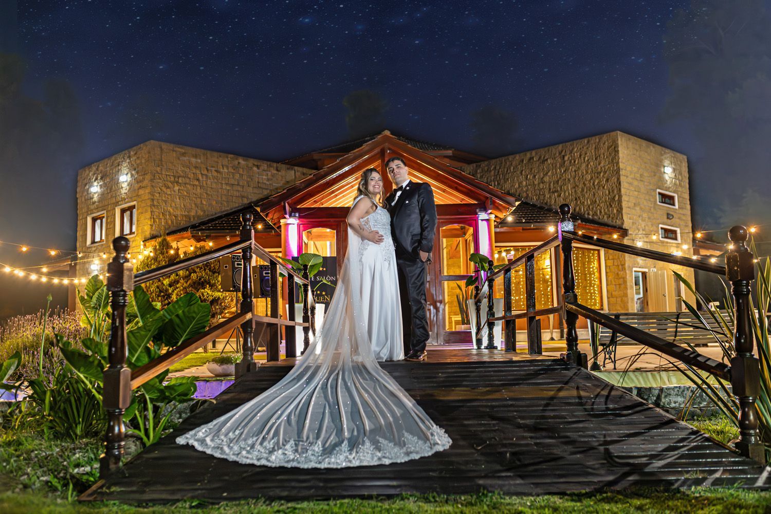 Pareja con traje de boda en un puente, posando frente a un edificio por la noche.