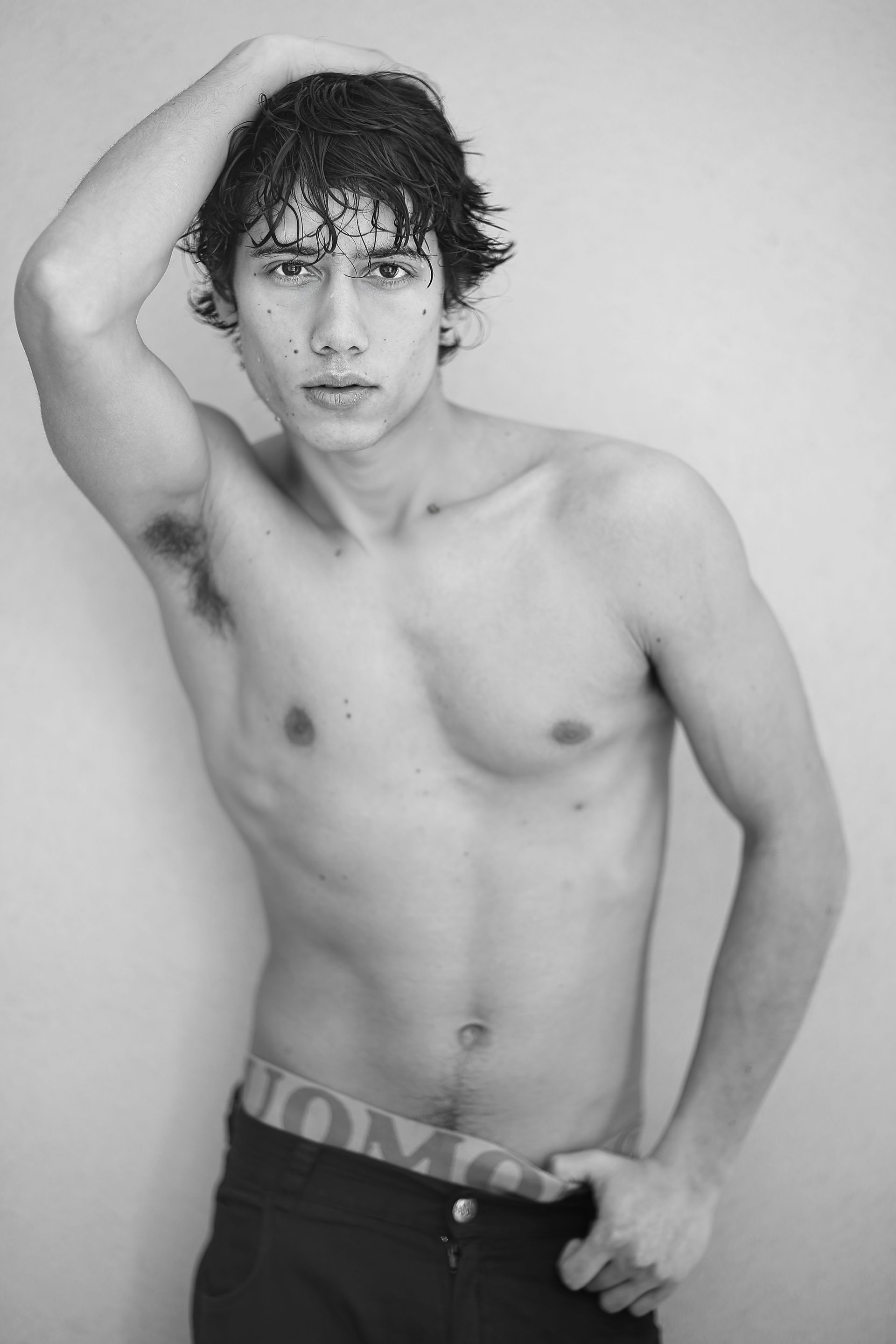 Hombre sin camisa, con la mano en el pelo, mirando a la cámara. Apoyado en una pared.