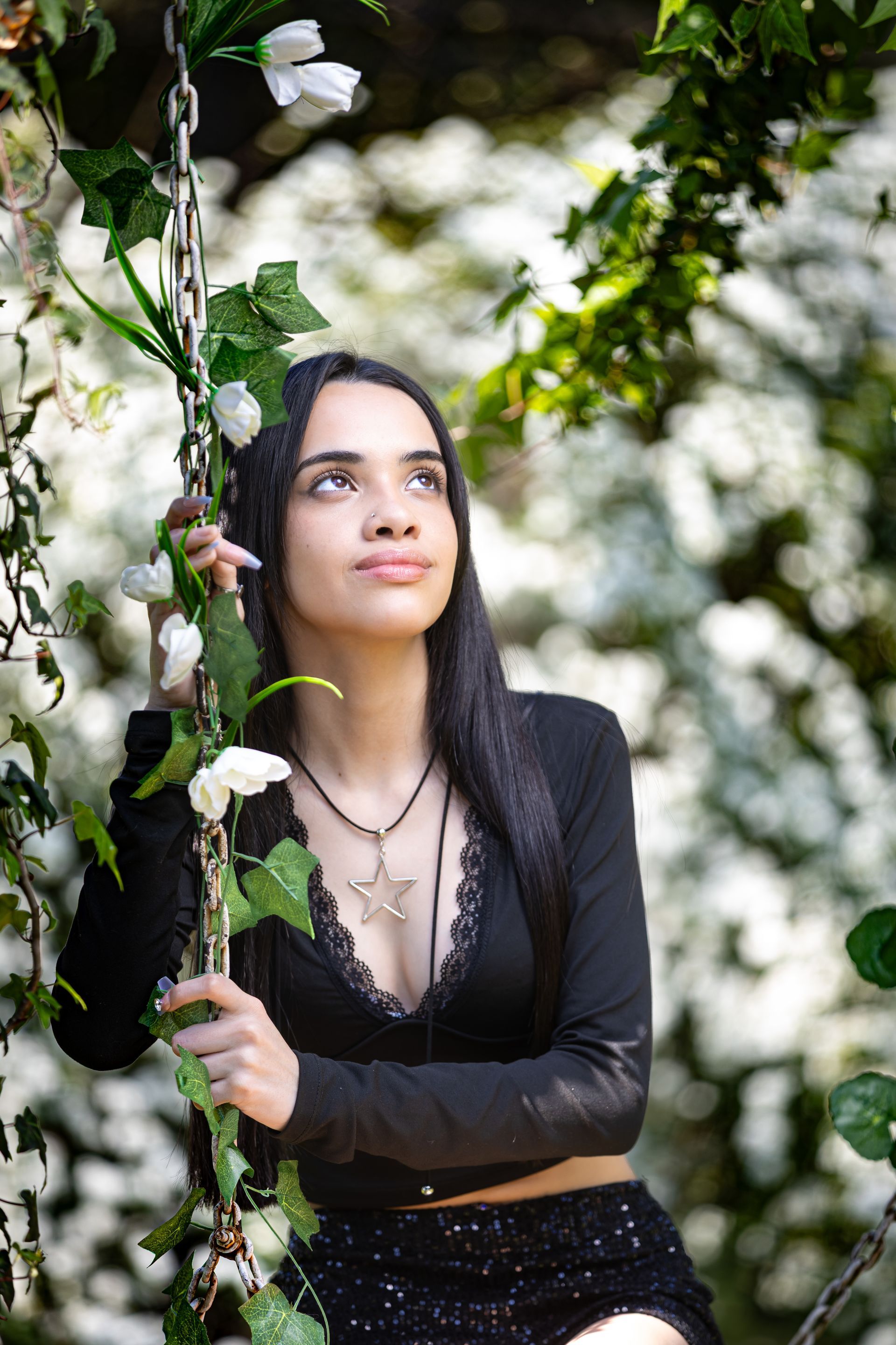 Mujer en un columpio, mirando hacia arriba, rodeada de flores blancas y enredaderas verdes. Lleva un top y una falda negros.