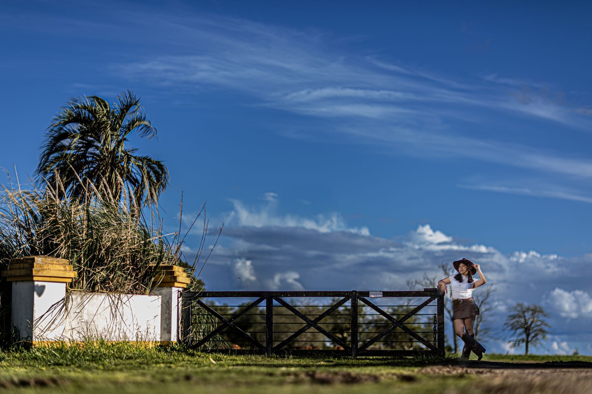 Una mujer con sombrero se apoya en una puerta, contra un cielo azul con nubes y una palmera cerca.
