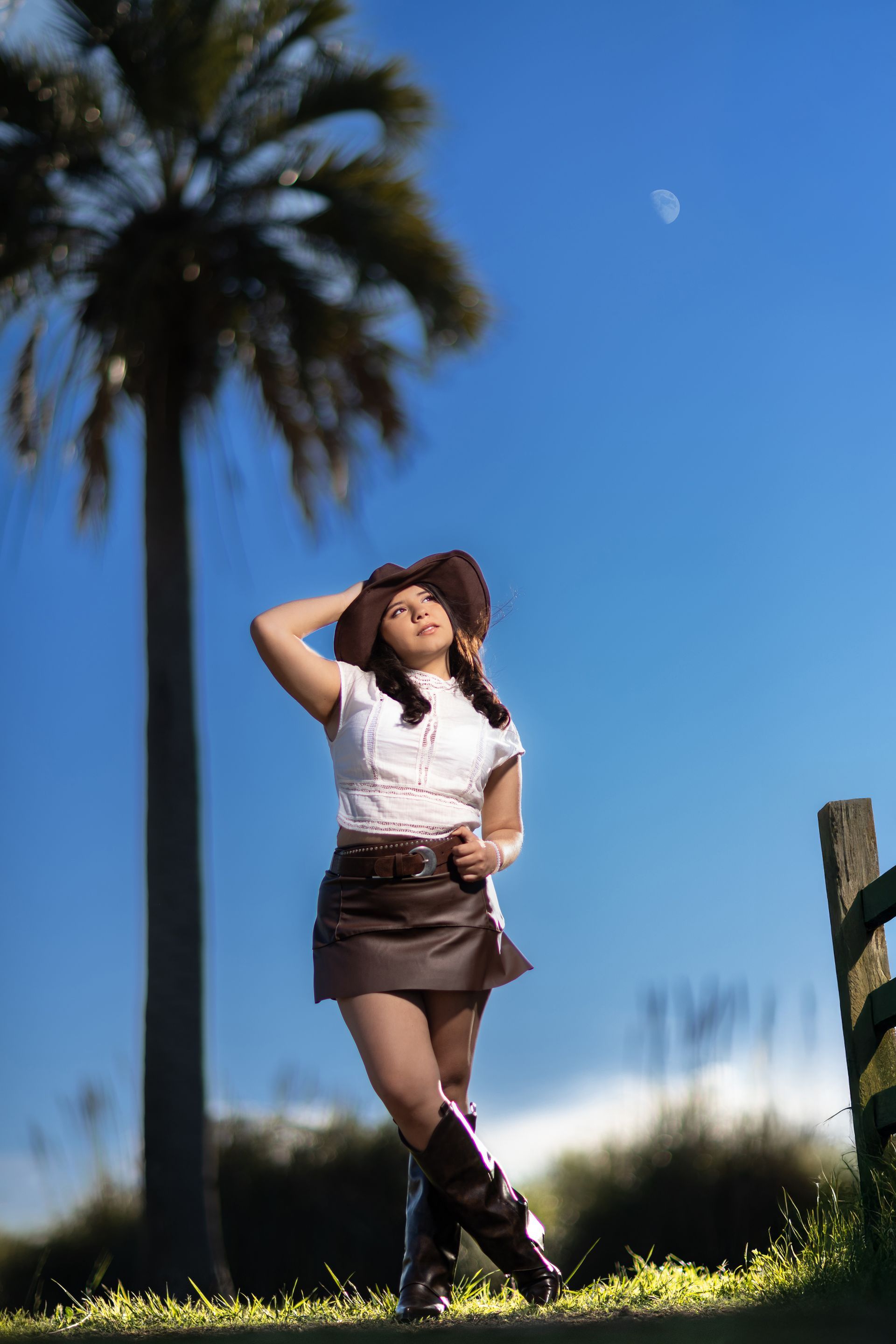Mujer con sombrero y botas de vaquero, de pie al aire libre, posando bajo una palmera con un cielo azul y una luna.