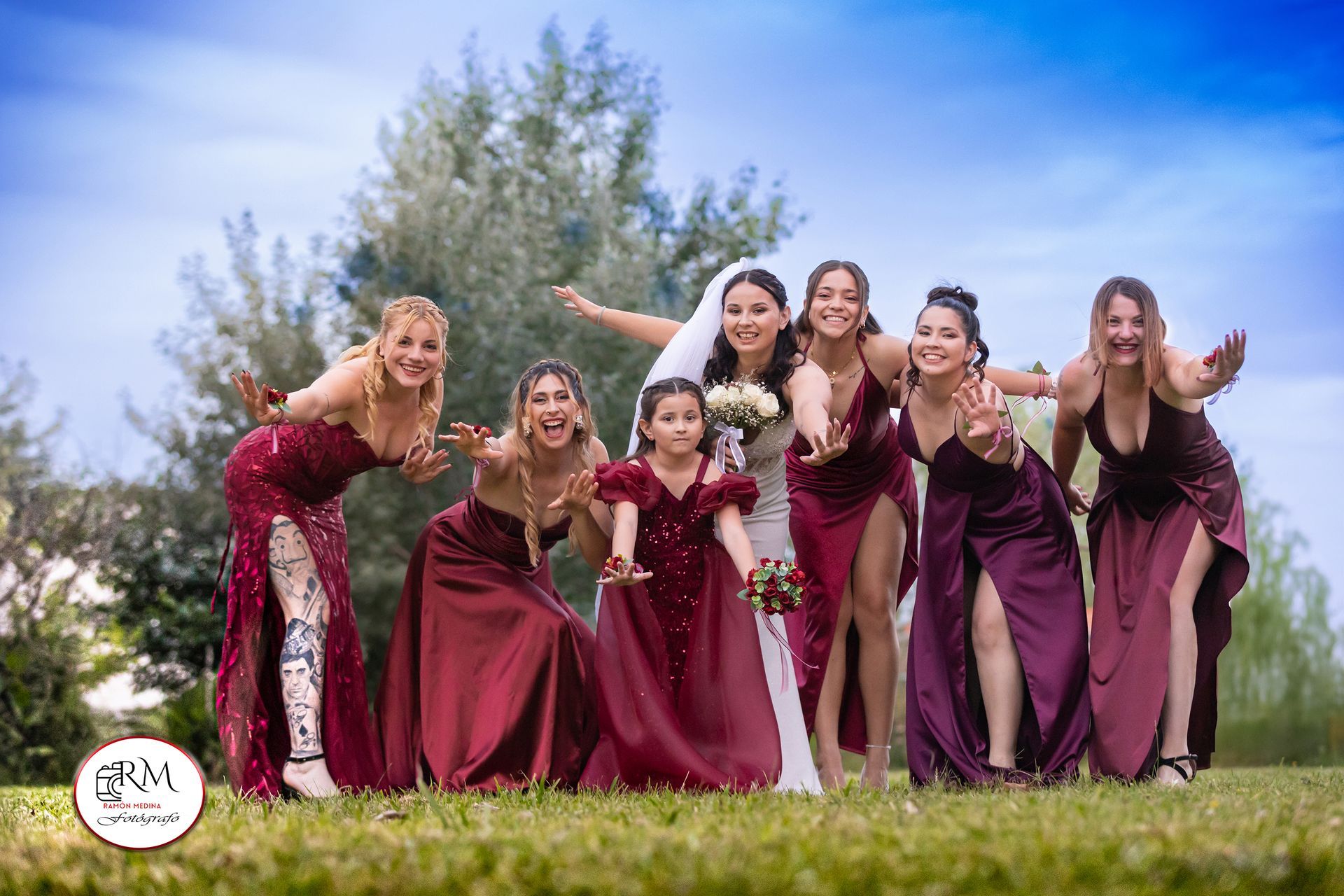 La novia y las damas de honor con vestidos color burdeos posan juguetonamente al aire libre.