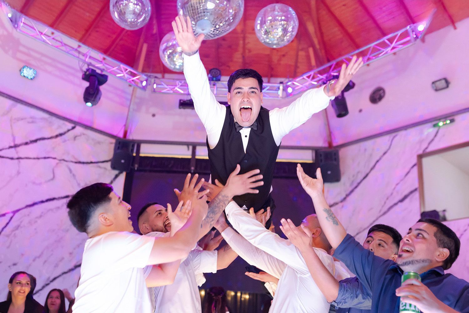 Un hombre sostenido en el aire por un grupo, celebrando en un lugar con bolas de discoteca e iluminación violeta.