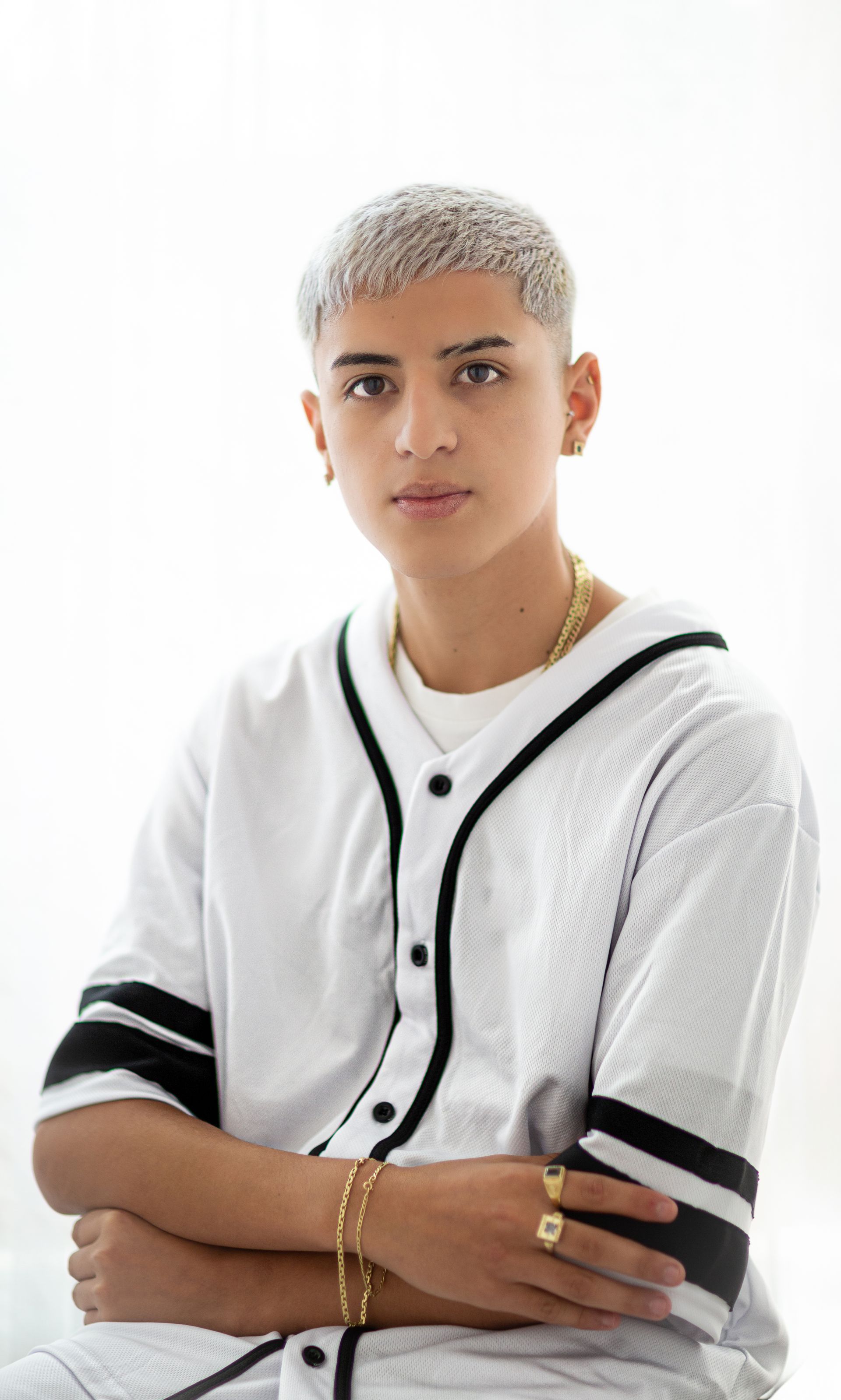 Persona joven con camiseta de béisbol blanca, cabello claro, joyas de oro, mirando al espectador.