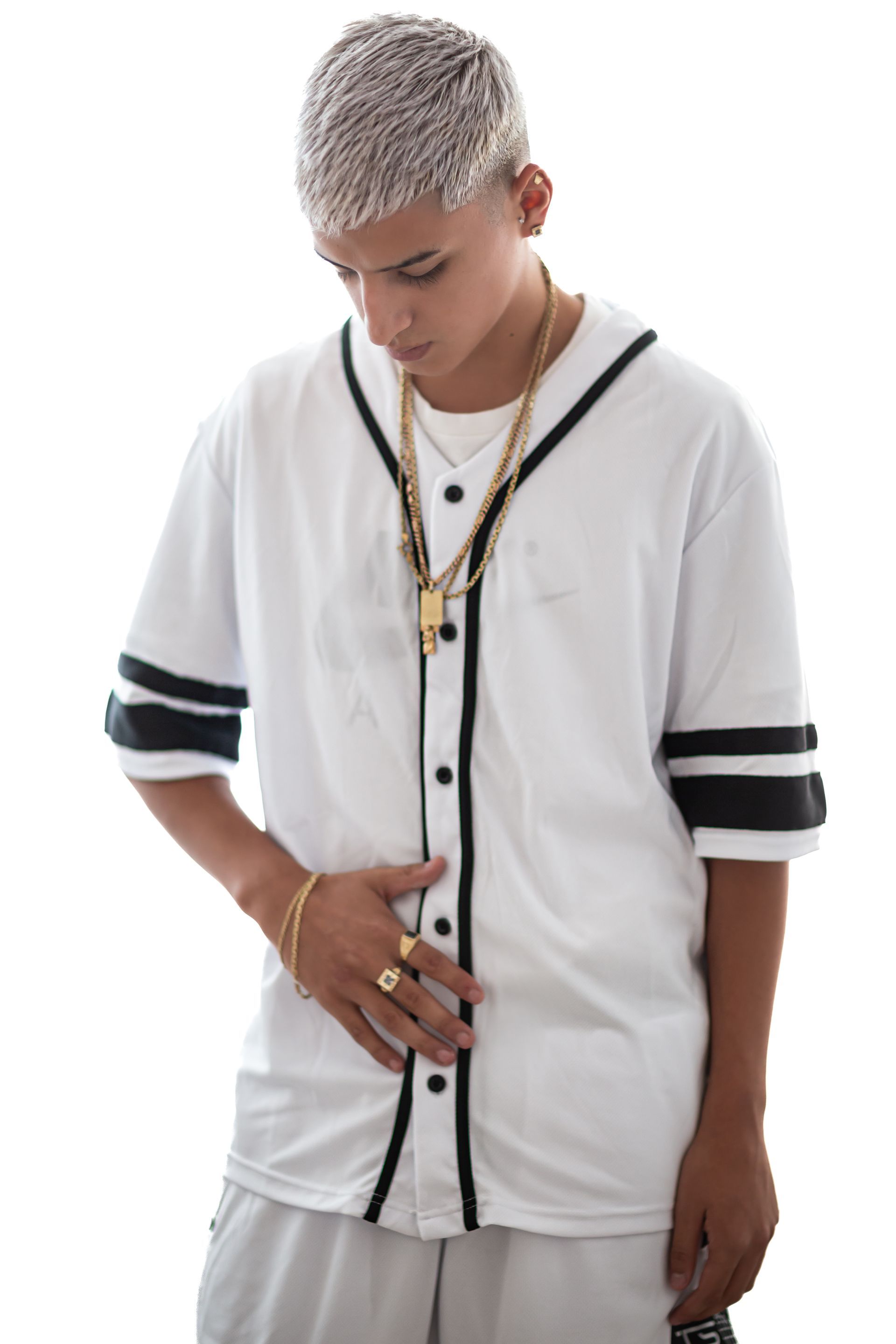 Persona joven con cabello platino, vistiendo una camiseta de béisbol blanca y joyas de oro, mirando hacia abajo.