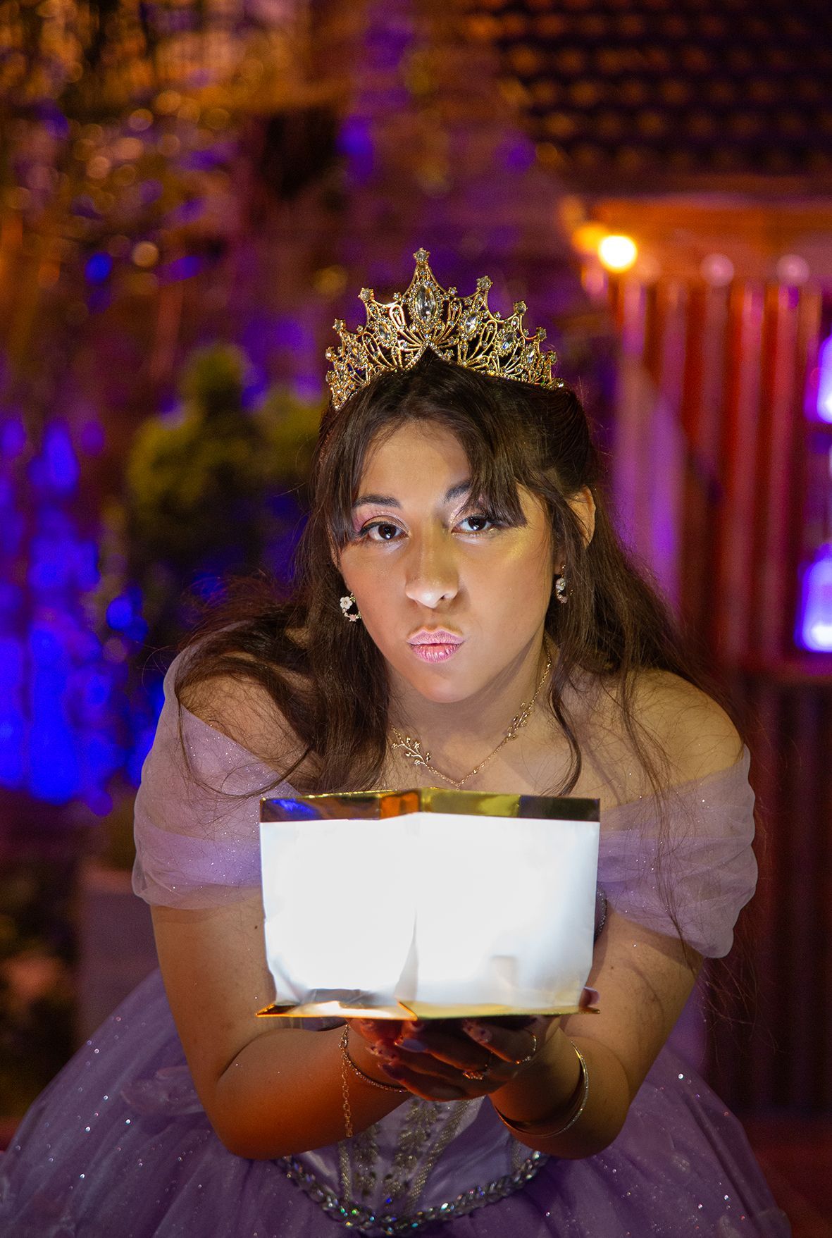 Mujer con corona, sosteniendo una caja encendida y lanzando un beso. Vestido morado, detalles dorados, ambientado de noche.