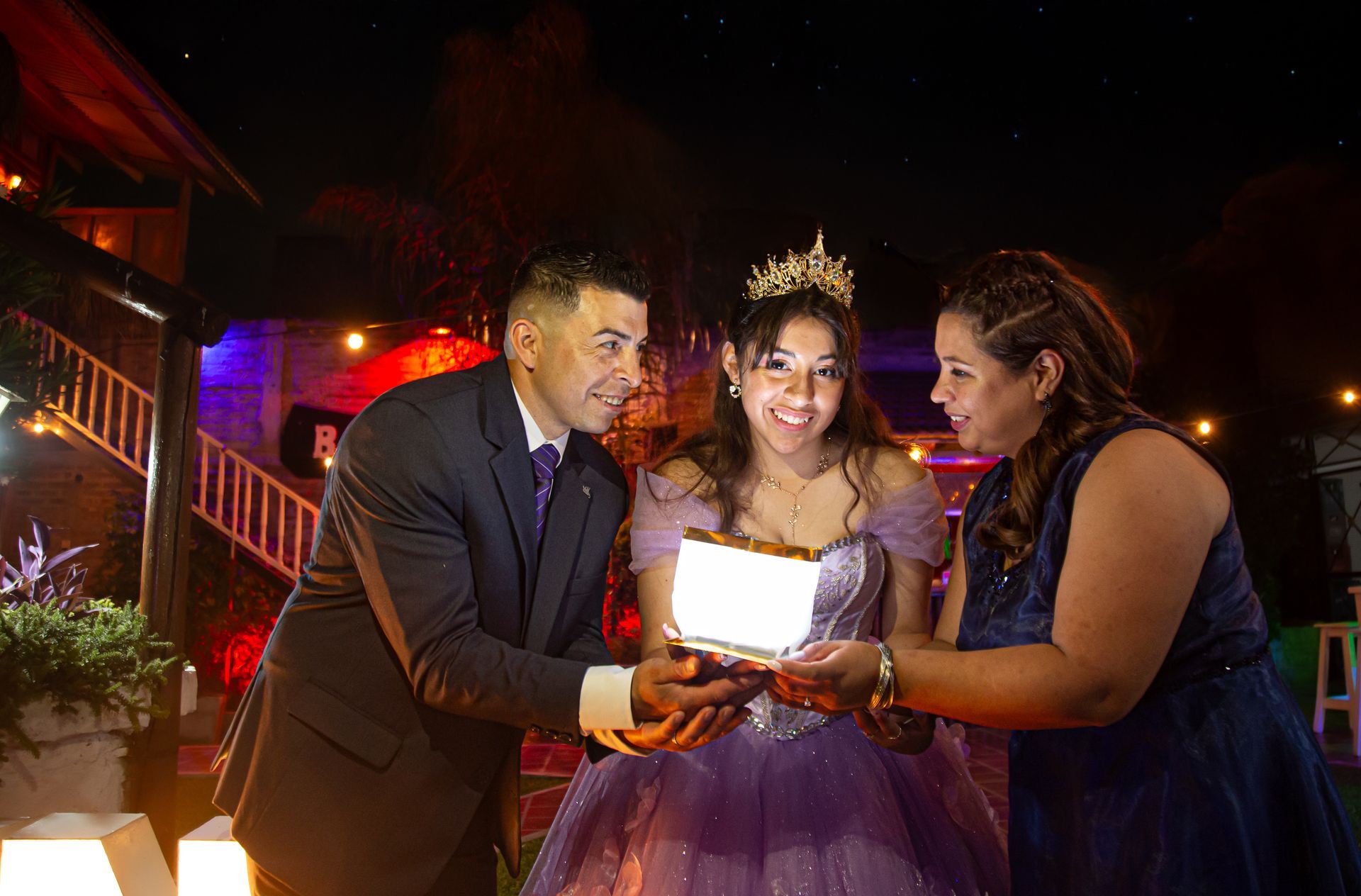 Una niña con una corona sonríe mientras sus padres sostienen una caja luminosa. Escena nocturna con luces y vestido morado.