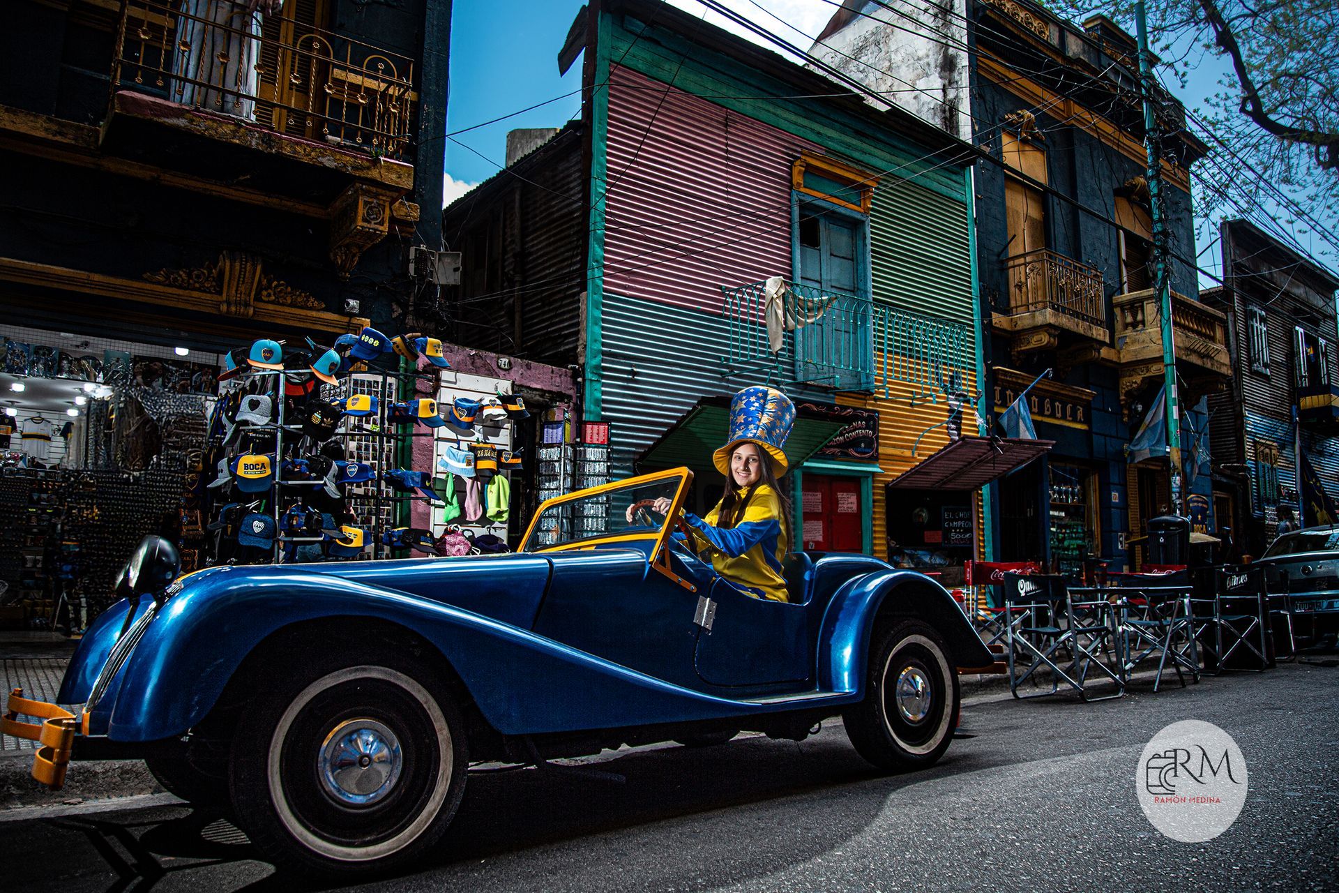 Un coche clásico azul en una calle de la ciudad con edificios coloridos. El conductor lleva sombrero.