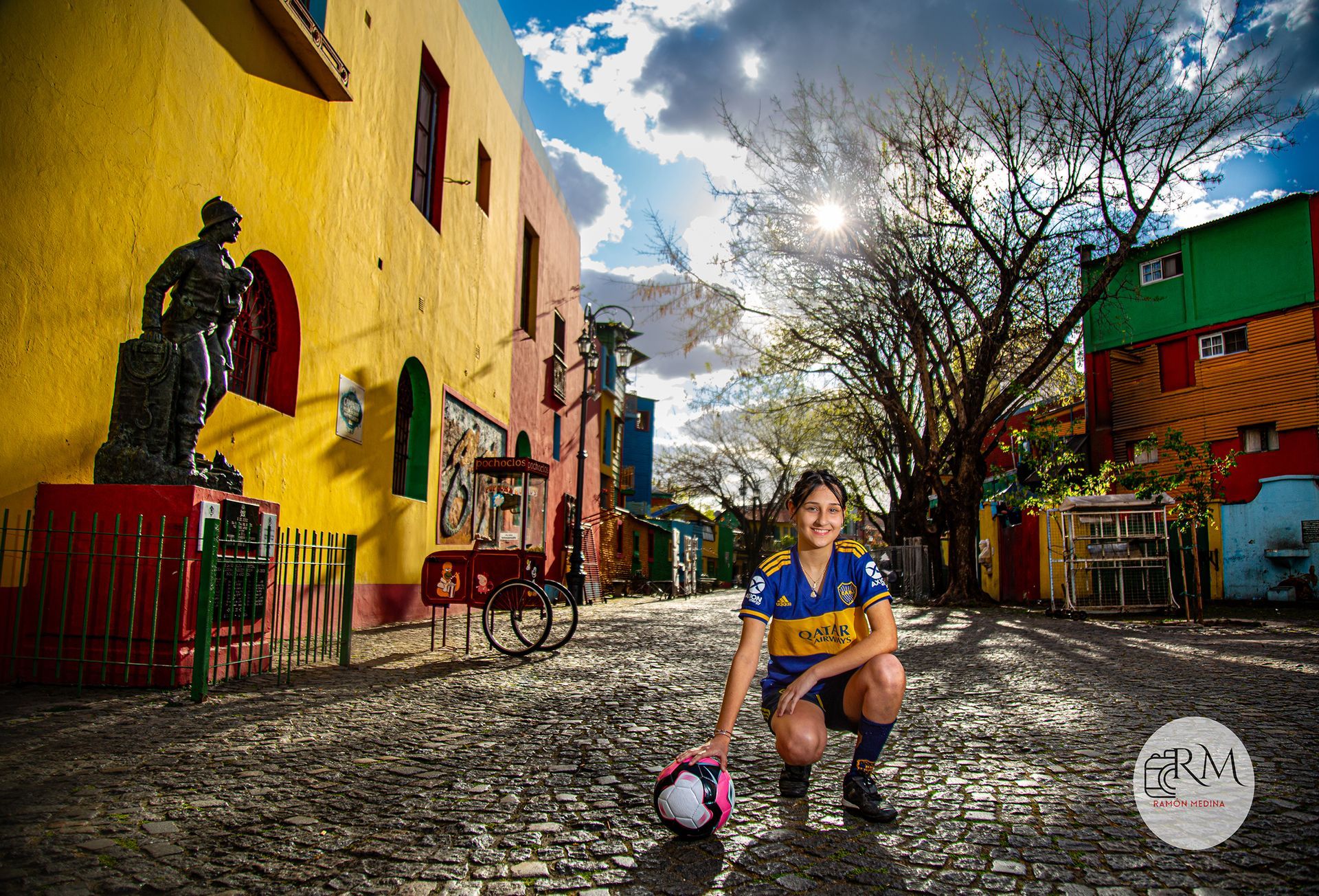 Una mujer con la camiseta de fútbol de Boca Juniors se arrodilla con un balón en una calle adoquinada