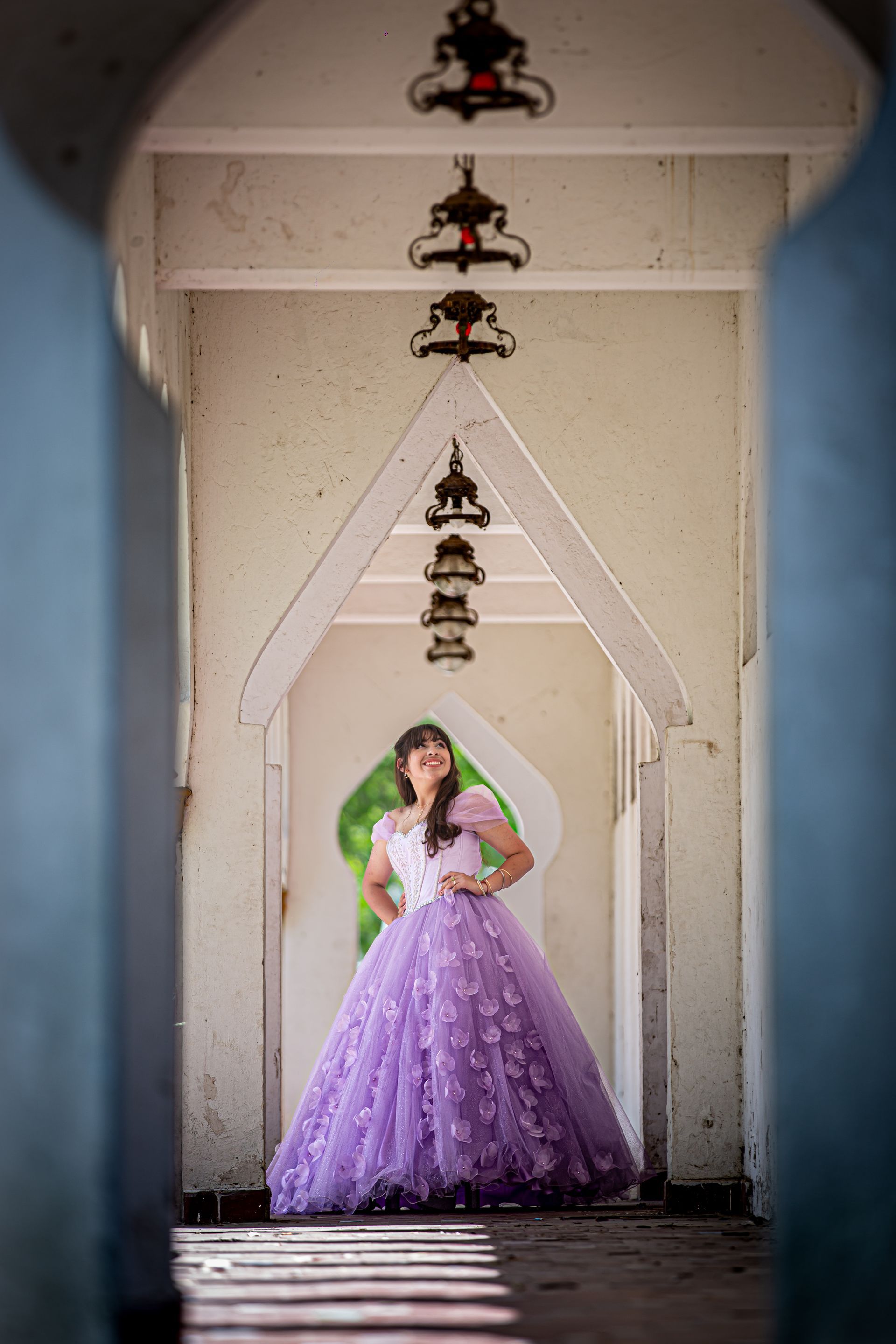 Una mujer con un vestido de gala color lavanda posa en un pasillo arqueado blanco con linternas colgantes ornamentadas.