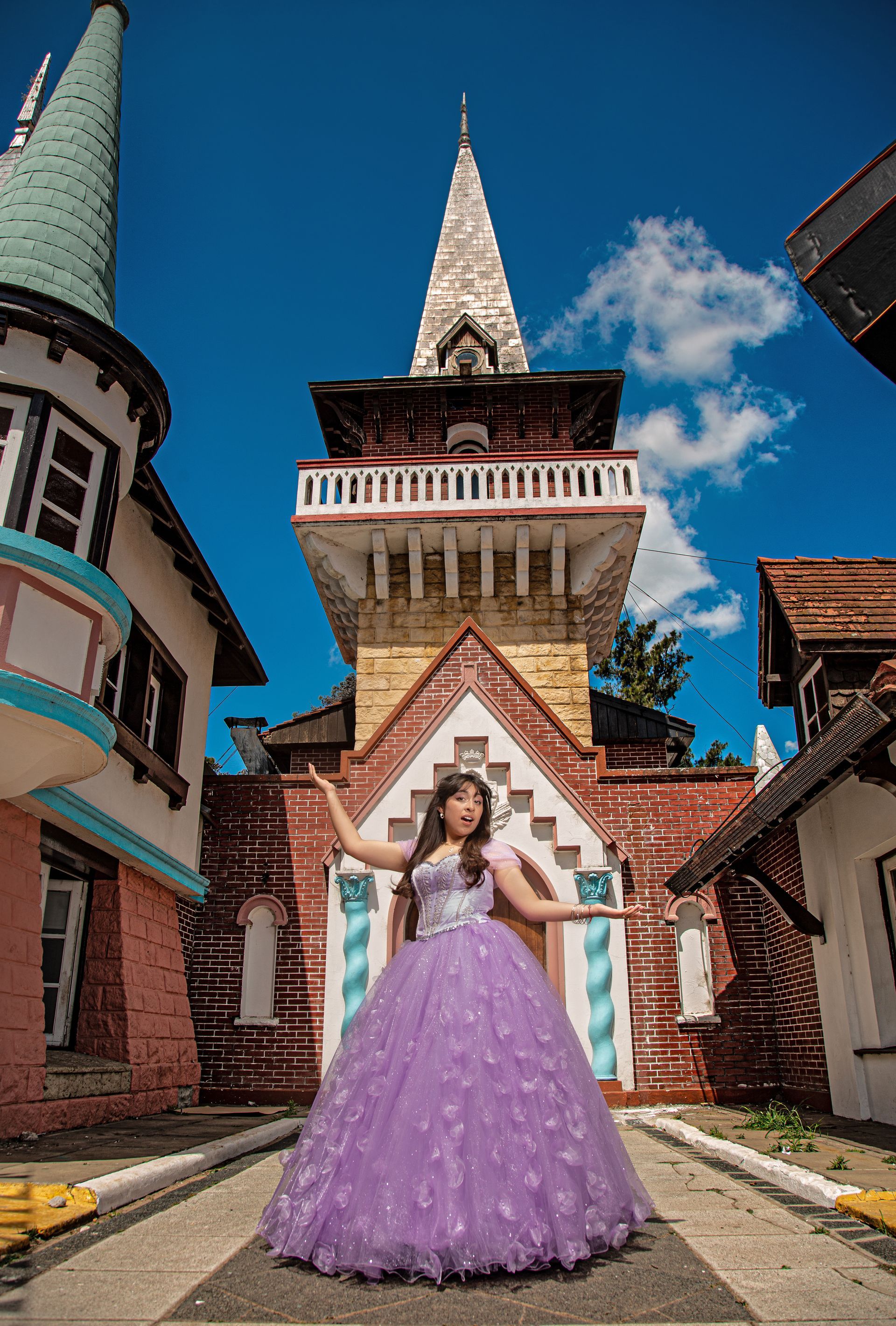 Una mujer con un vestido morado posa frente a una torre de ladrillos y edificios coloridos en un día soleado.