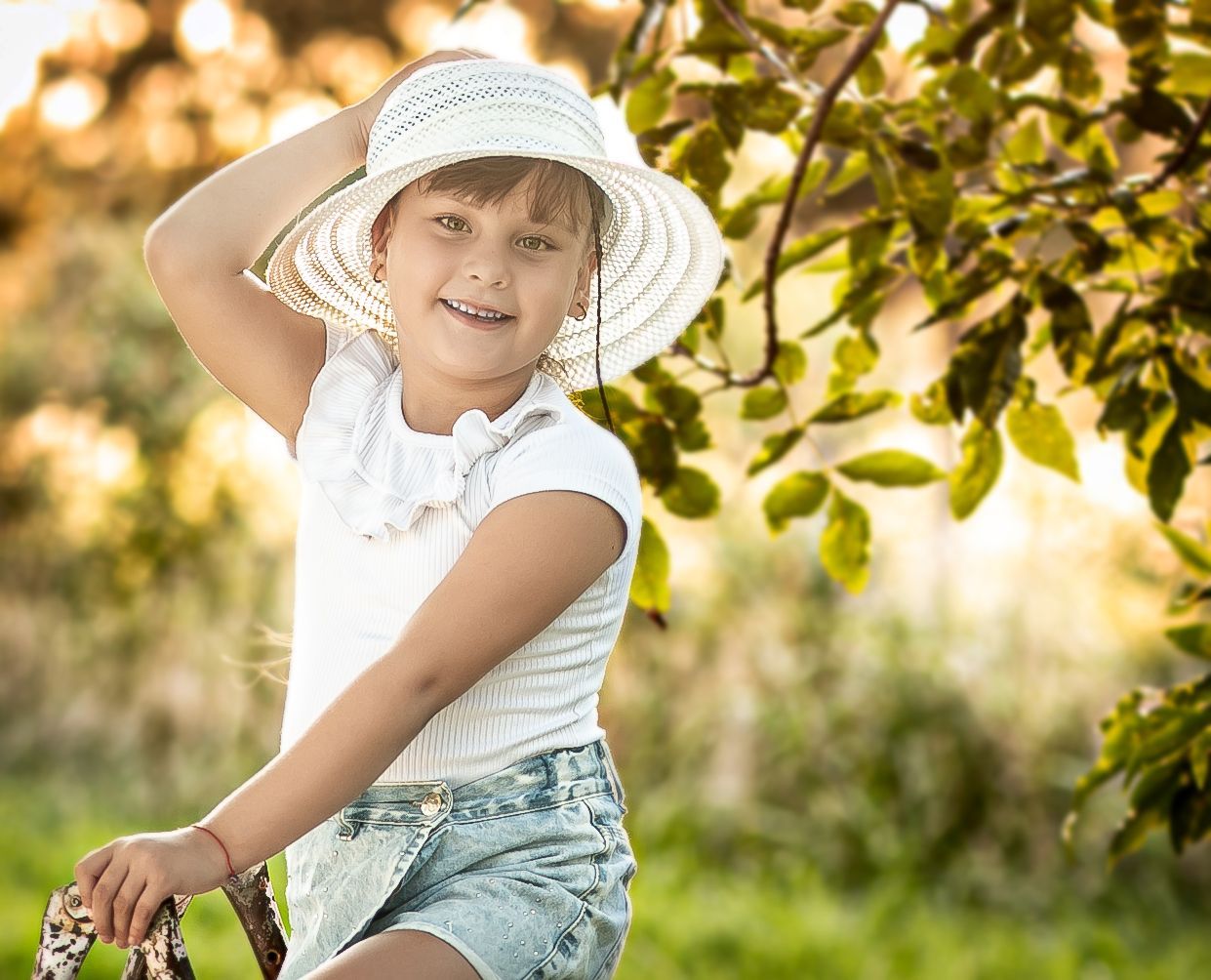 Niña con sombrero y camisa blanca, sonriendo, posando junto a un árbol en un entorno soleado al aire libre.