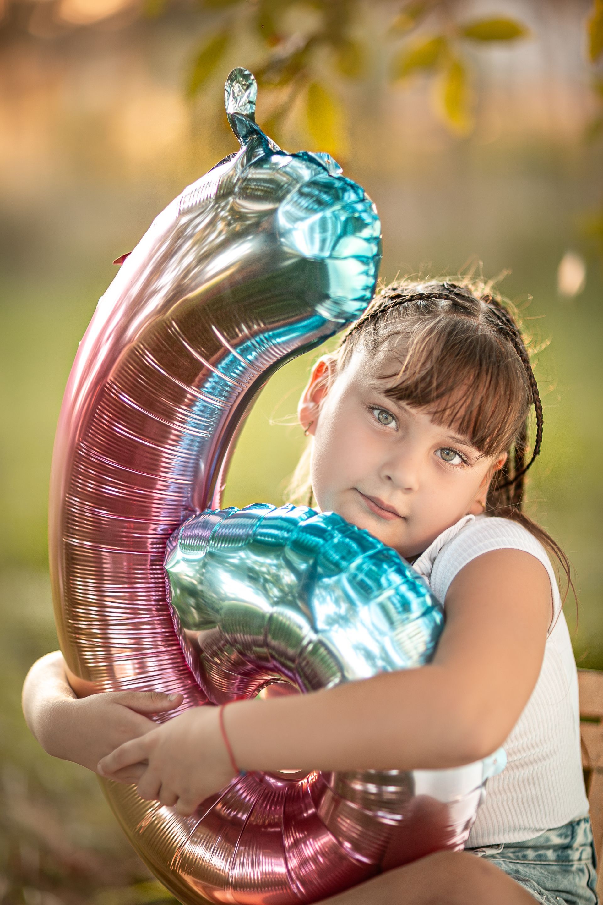 Niña abrazando un globo número 6 al aire libre, sonriendo, con colores azul, rosa y plata.
