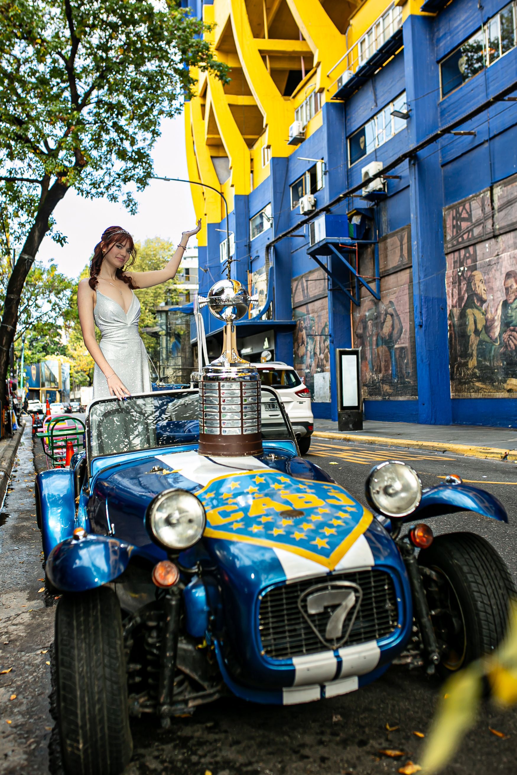 Una mujer vestida de plata está parada en un auto deportivo azul, sosteniendo un trofeo frente al estadio de Boca Juniors.