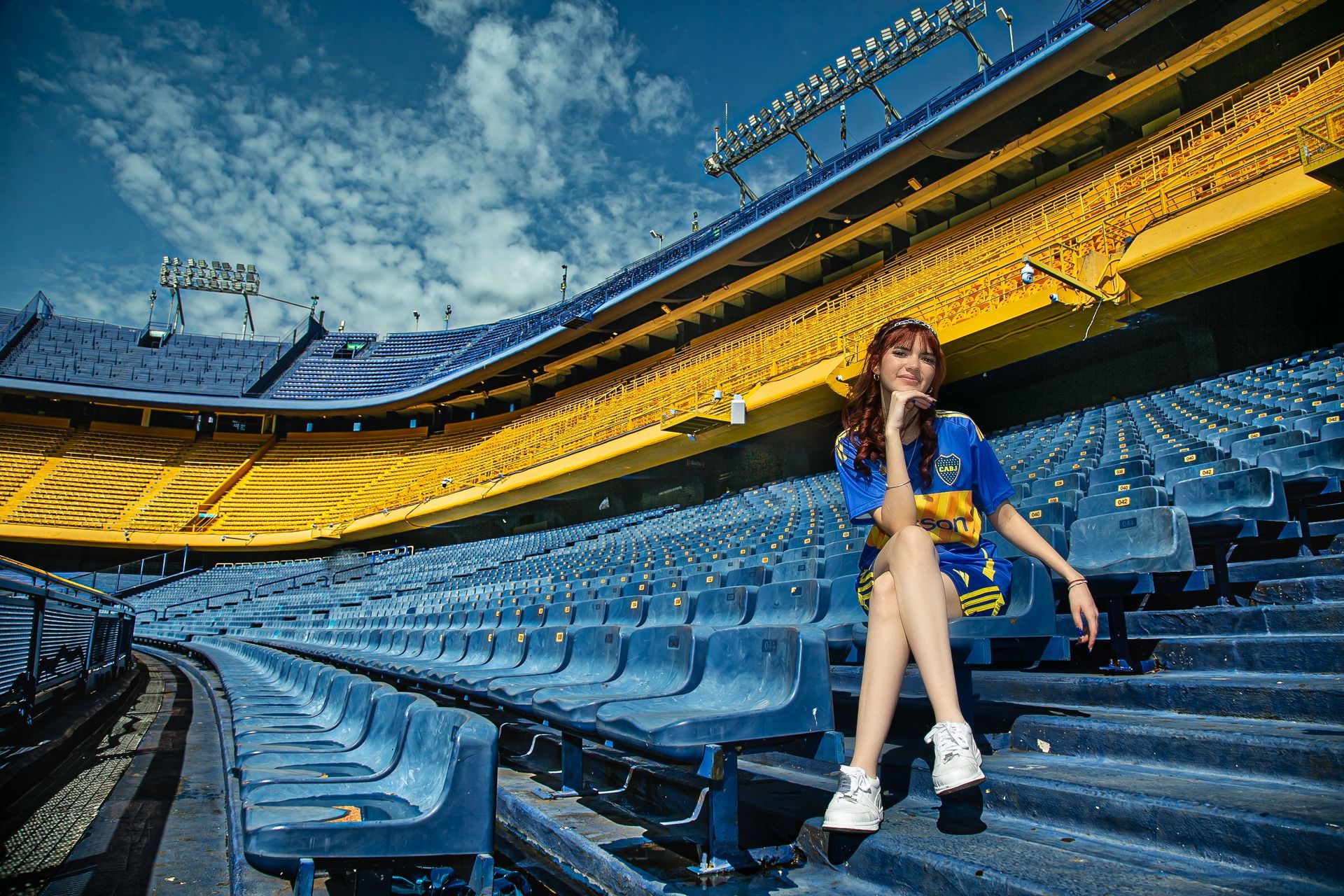 Mujer sentada en el estadio de Boca Juniors, con los colores del equipo. Asientos azules, paredes amarillas y cielo azul.