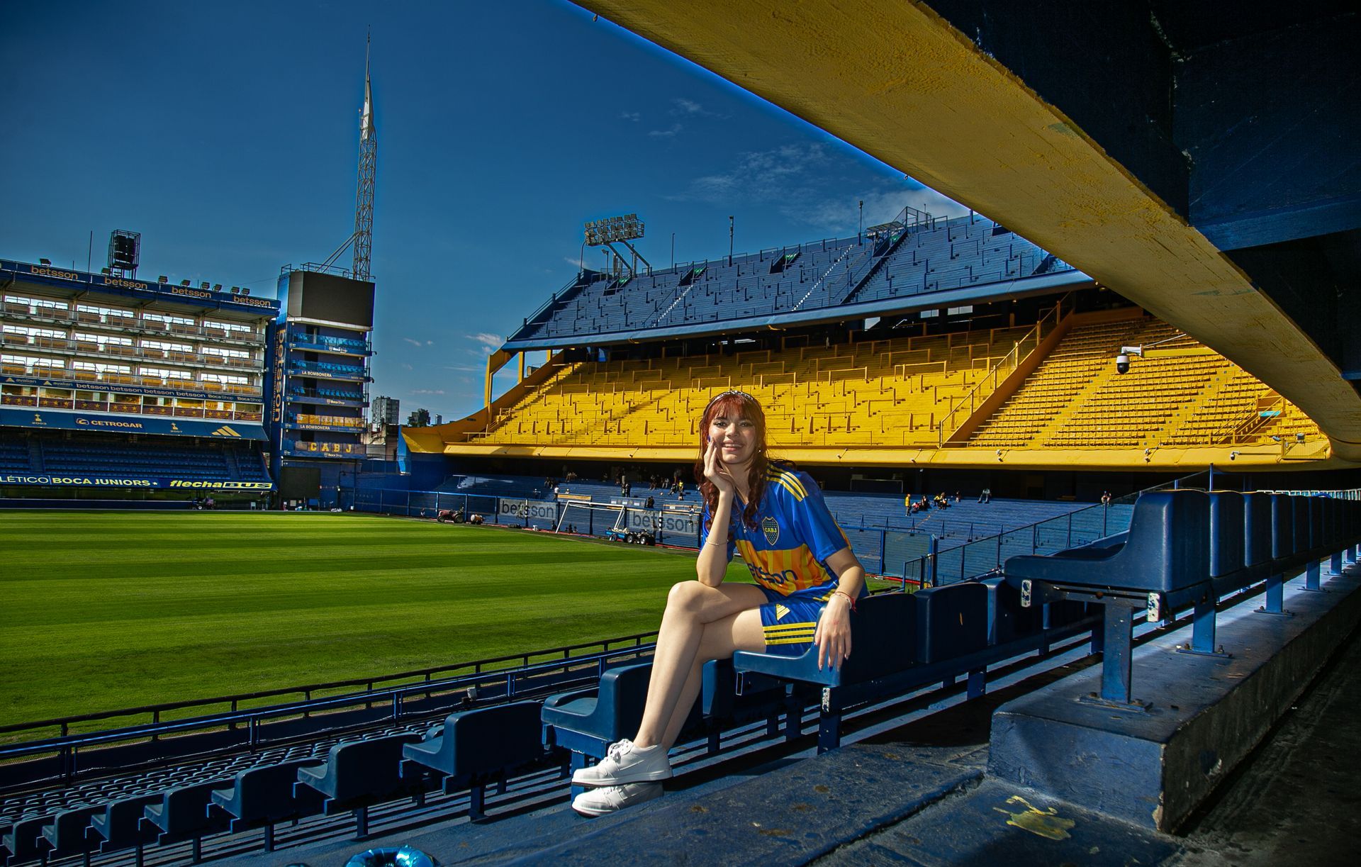 Una mujer con la camiseta de Boca Juniors sonríe en el estadio La Bombonera, con asientos y cancha azul y amarillo visibles.