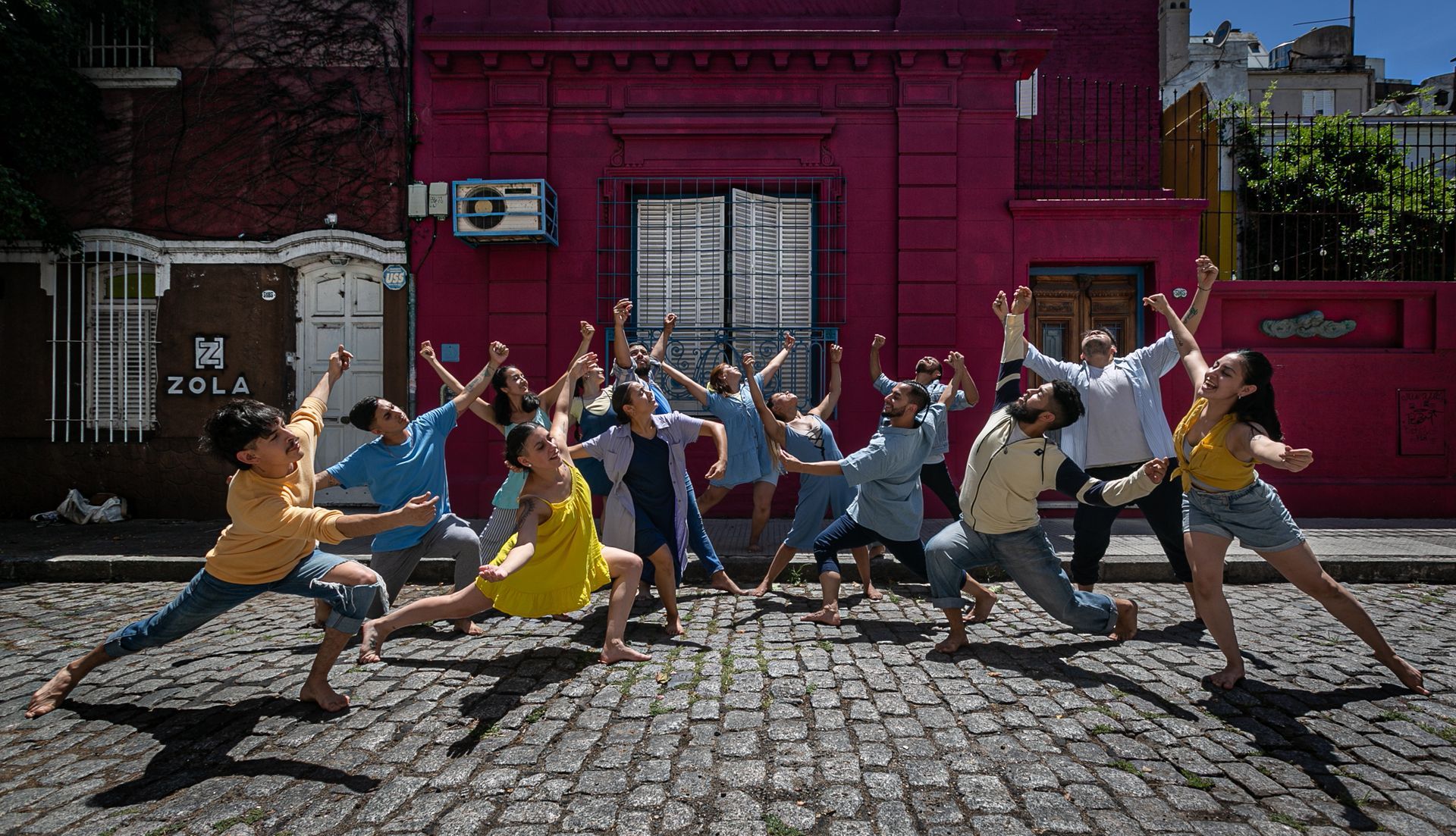 Grupo de personas bailando frente a un edificio magenta en una calle adoquinada.