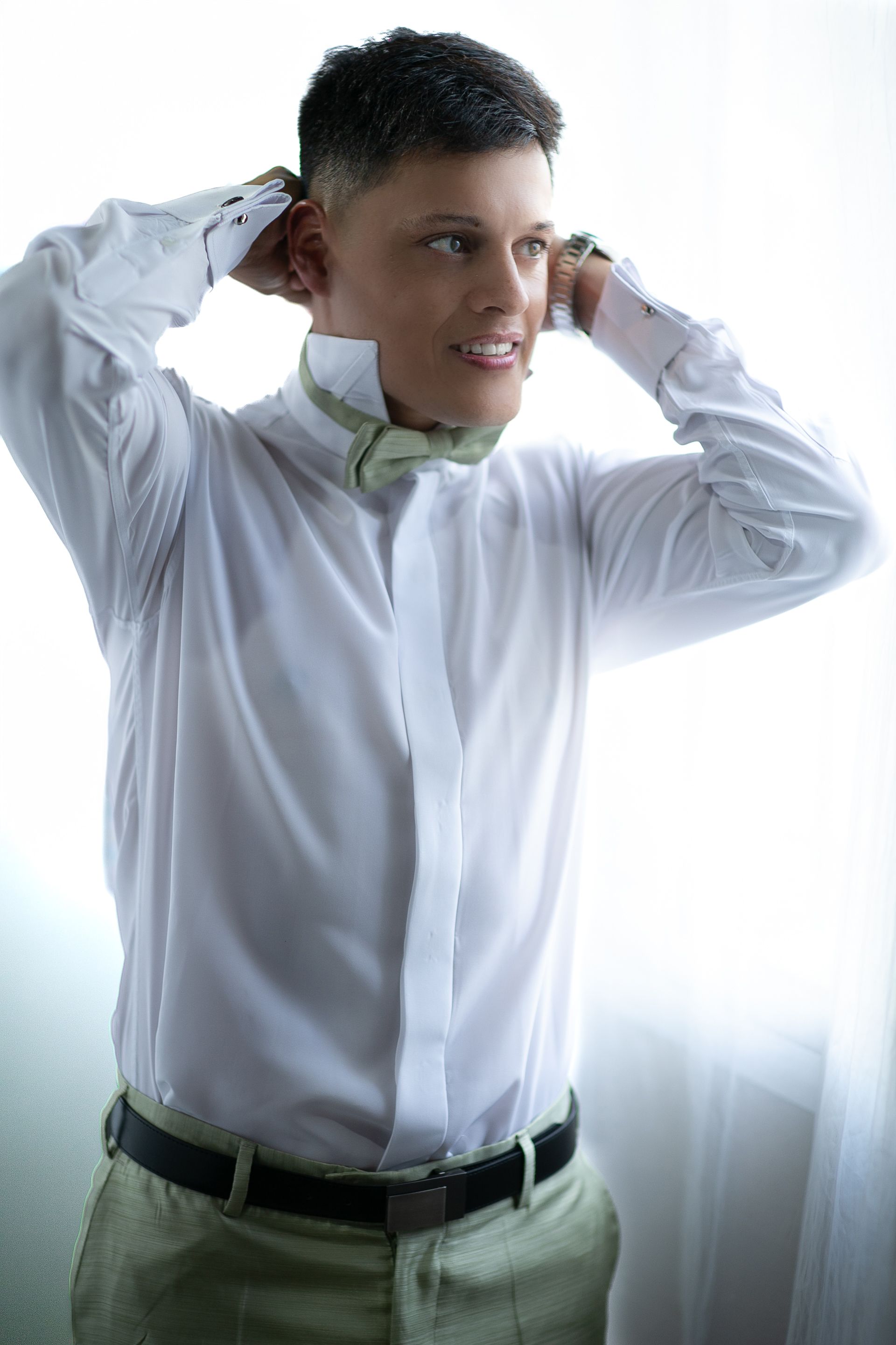Hombre con camisa blanca y pajarita ajustándose el cuello. Pantalones verdes y cinturón negro. Luz natural.