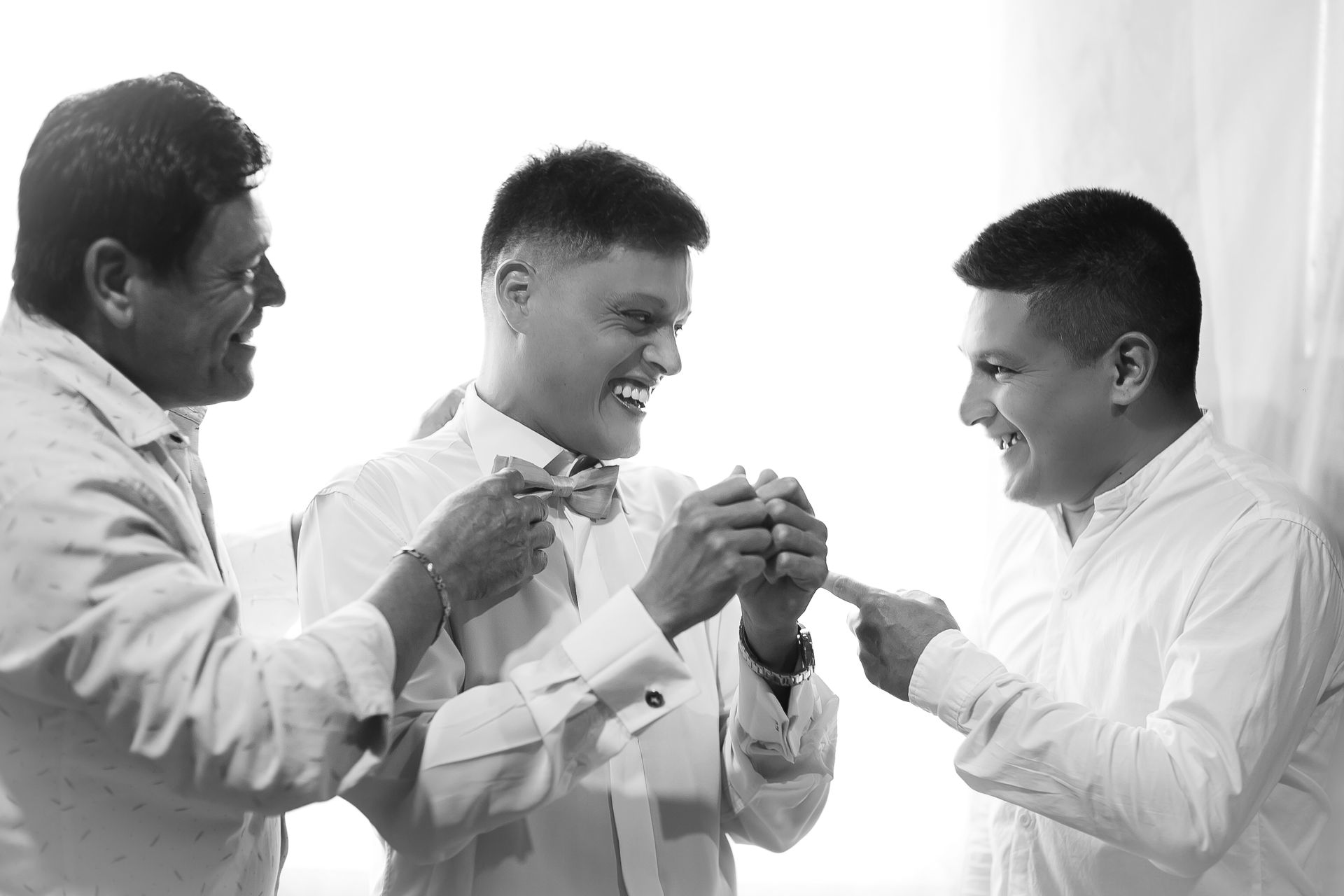 Tres hombres con camisa blanca, uno ajustándose la pajarita, todos riendo. Interior luminoso.