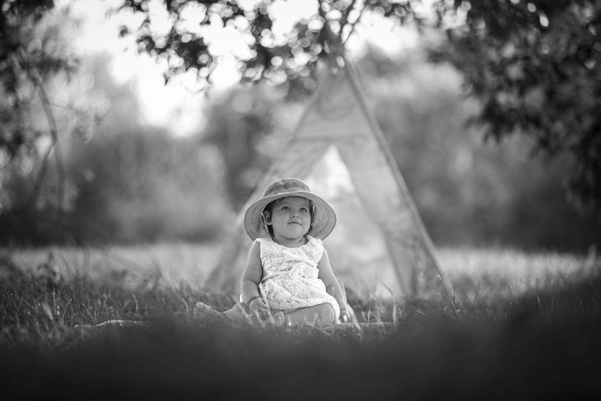 Un niño con sombrero para el sol se sienta en la hierba alta cerca de un tipi, con árboles al fondo. Foto en blanco y negro.