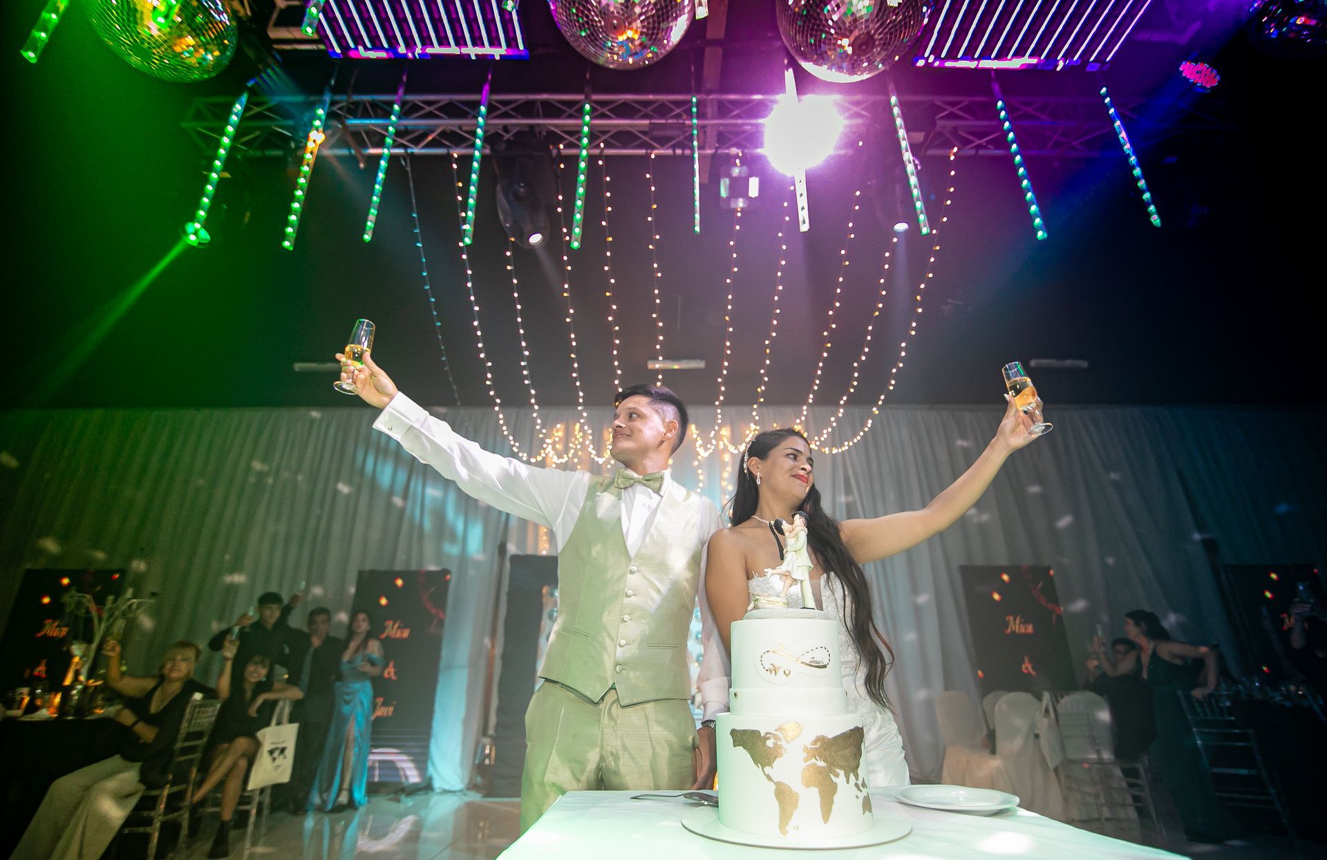 Pareja brindando con champán junto a una tarta nupcial; pista de baile iluminada con bolas de discoteca.