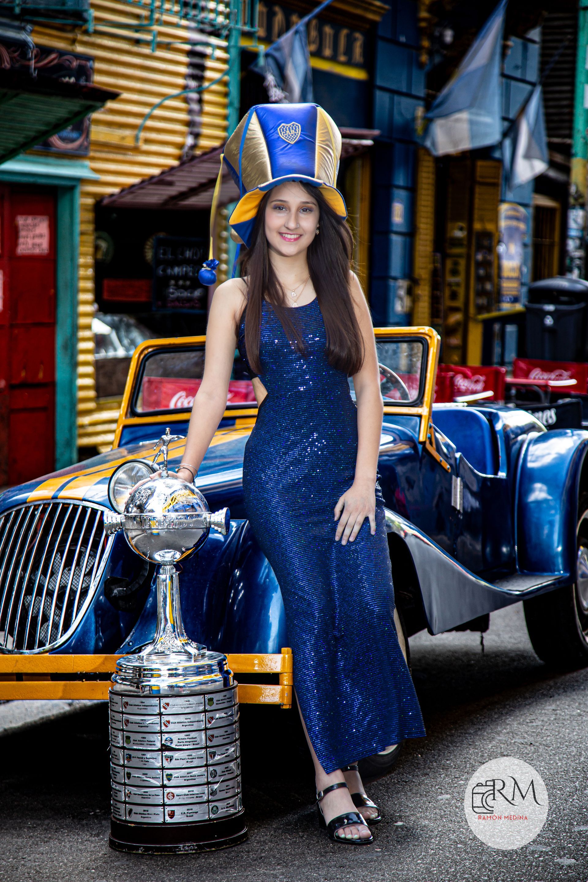 Mujer con vestido azul, trofeo y sombrero temático junto a un coche clásico azul.