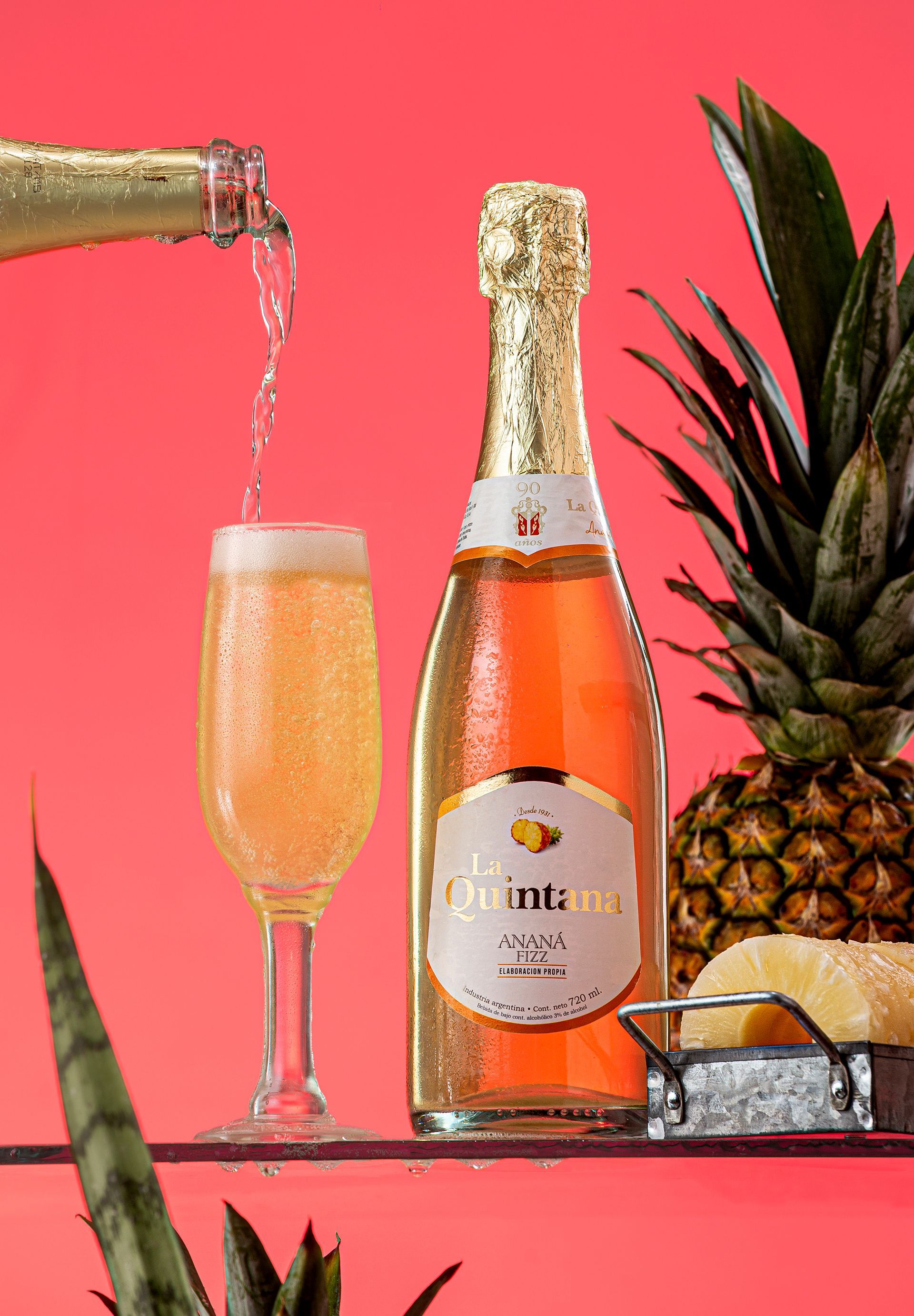 Botella de vino espumoso vertiéndose en una copa de champán; piña en un estante sobre un fondo rosa.