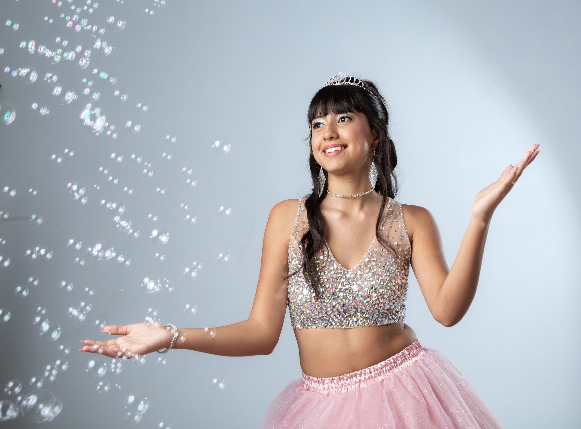 Adolescente vestida de gala con tiara, sonriendo con los brazos extendidos y burbujas flotando a su alrededor.