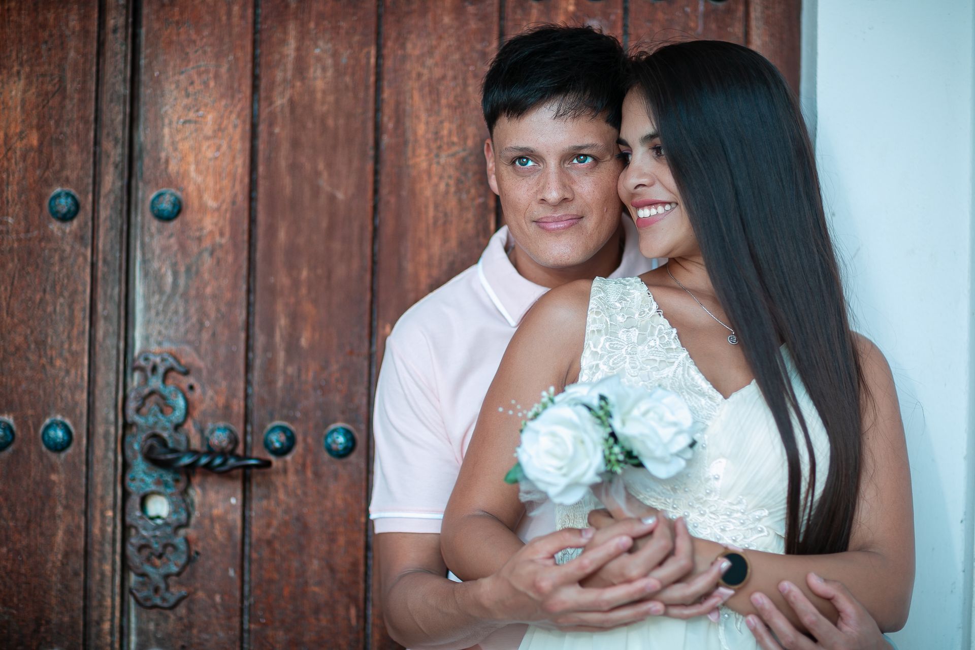 Pareja abrazándose, mujer sosteniendo flores, sonriendo, cerca de una puerta de madera.